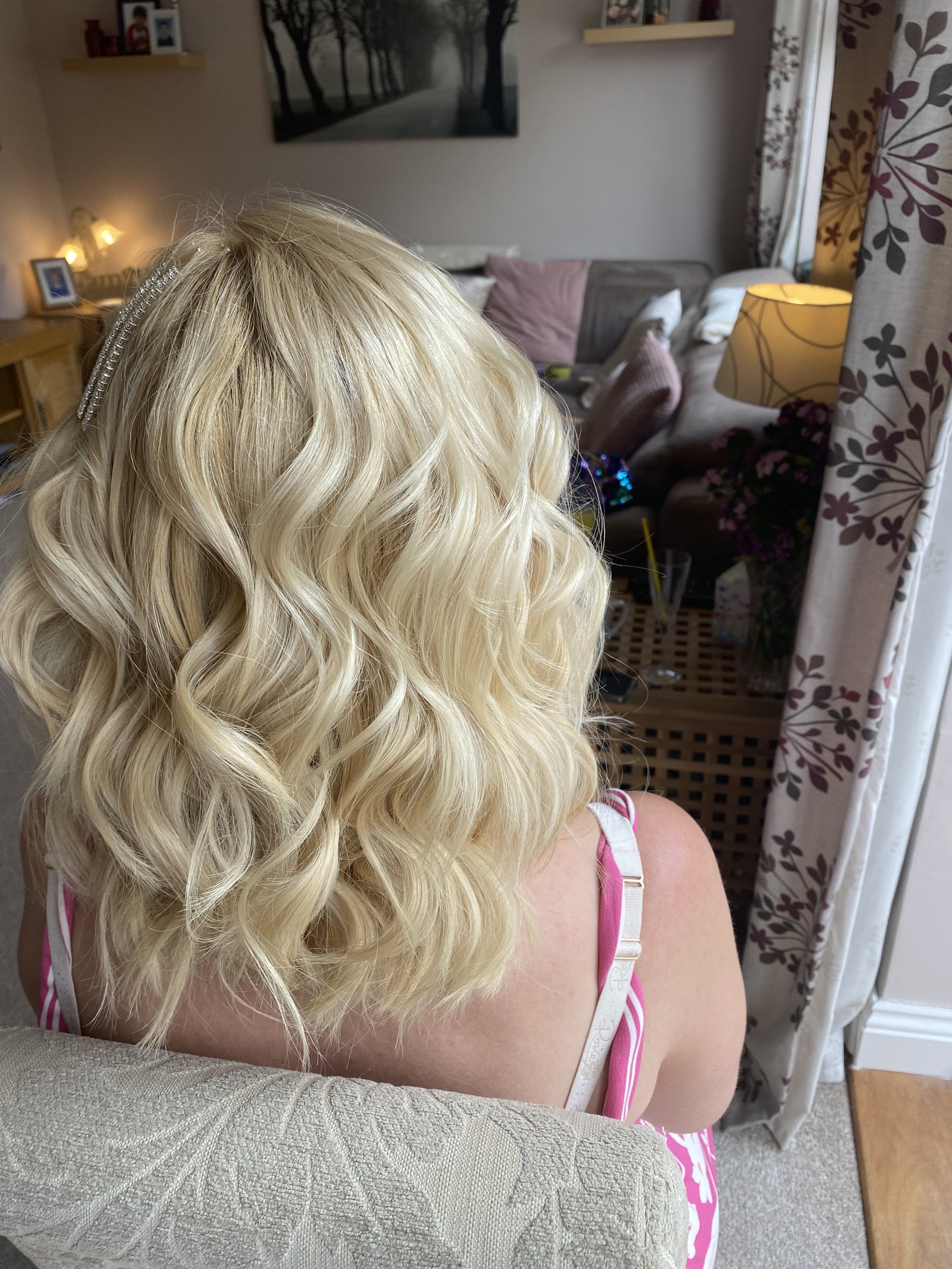 Rear view of a woman with long, blonde, curly hair, wearing a pink and white striped tank top, sitting on a beige couch in a living room.
