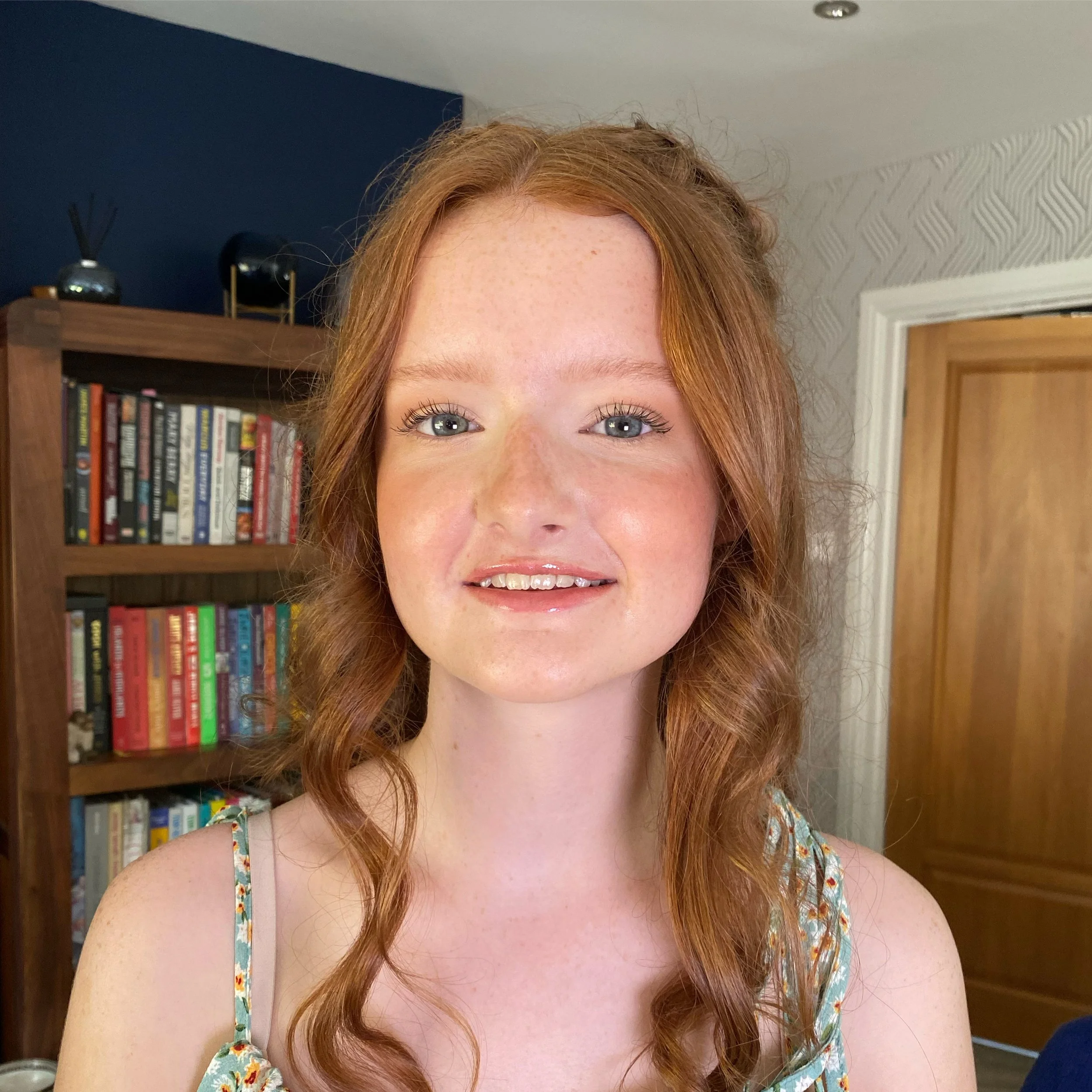 A woman with red hair, blue eyes, and freckles smiling in front of a bookshelf in a room with a wooden door.