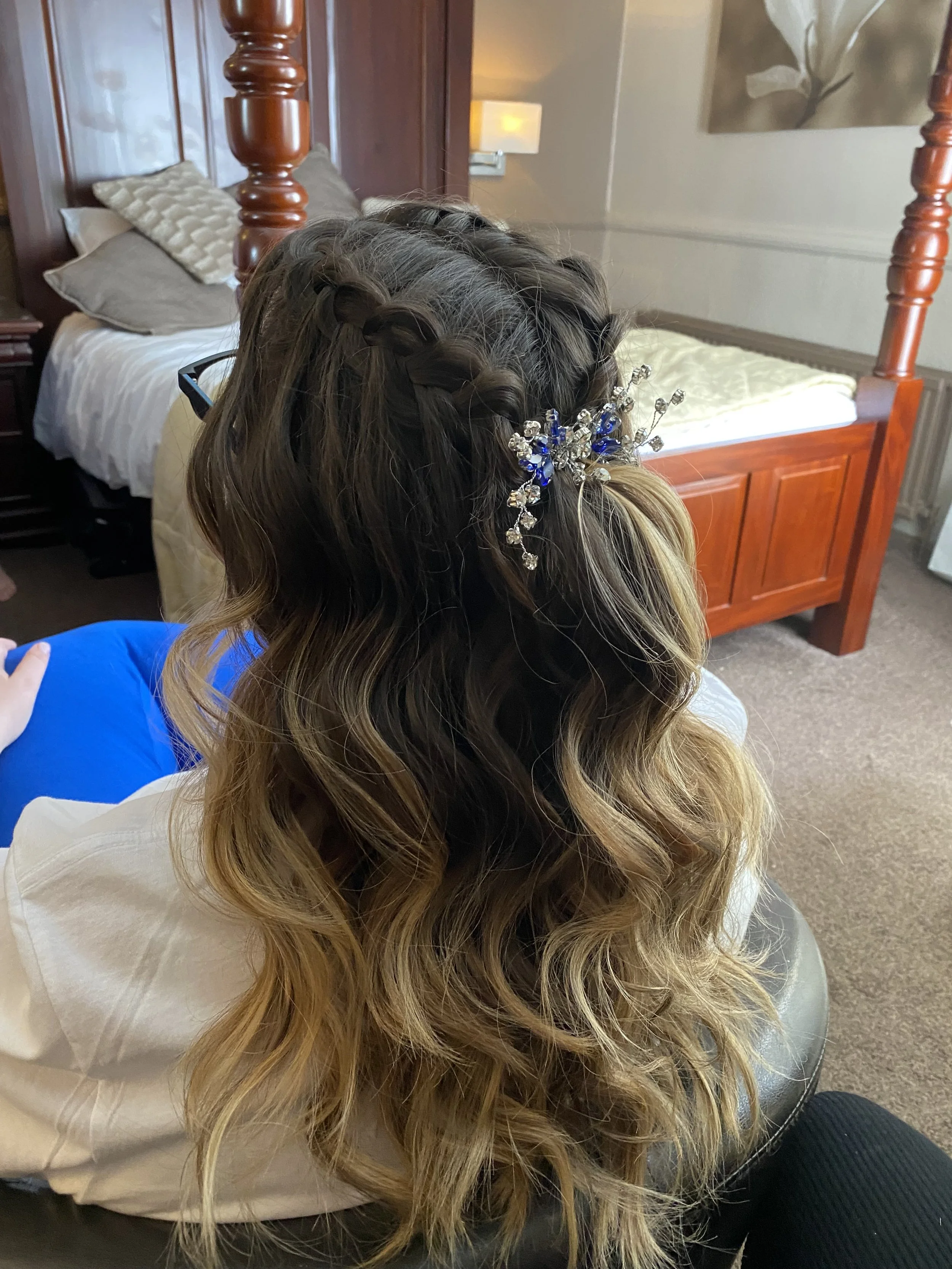 A woman sitting with her long, wavy hair styled in a braid with a decorative silver and blue hairpiece, in a bedroom with a bed, pillows, and wall art.