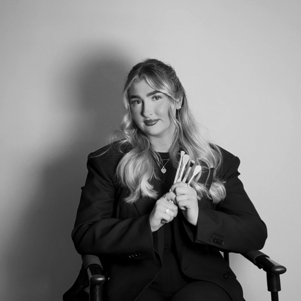 A woman with long wavy hair, wearing a black blazer, sitting in a chair, holding makeup brushes, smiling, in a black and white portrait.