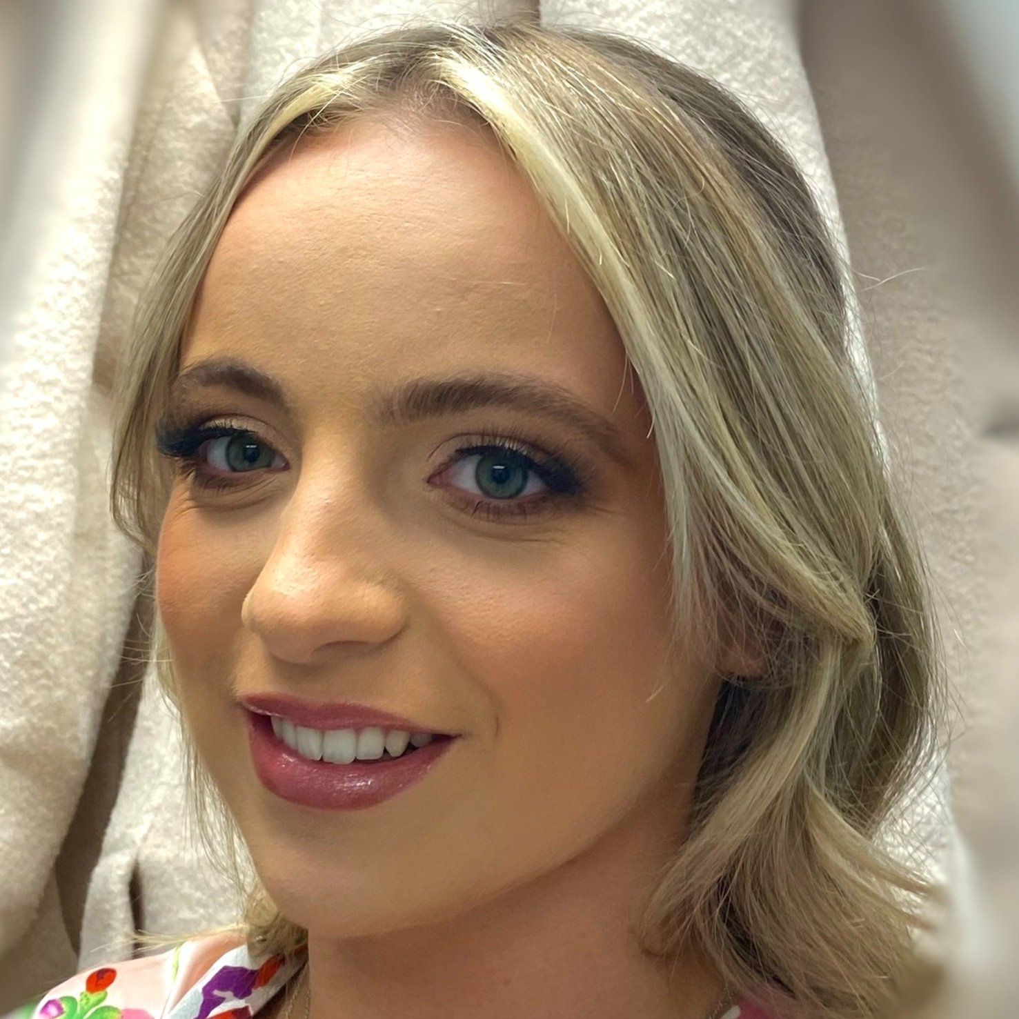 A smiling woman with blonde hair and blue eyes, wearing makeup, in front of a beige background.