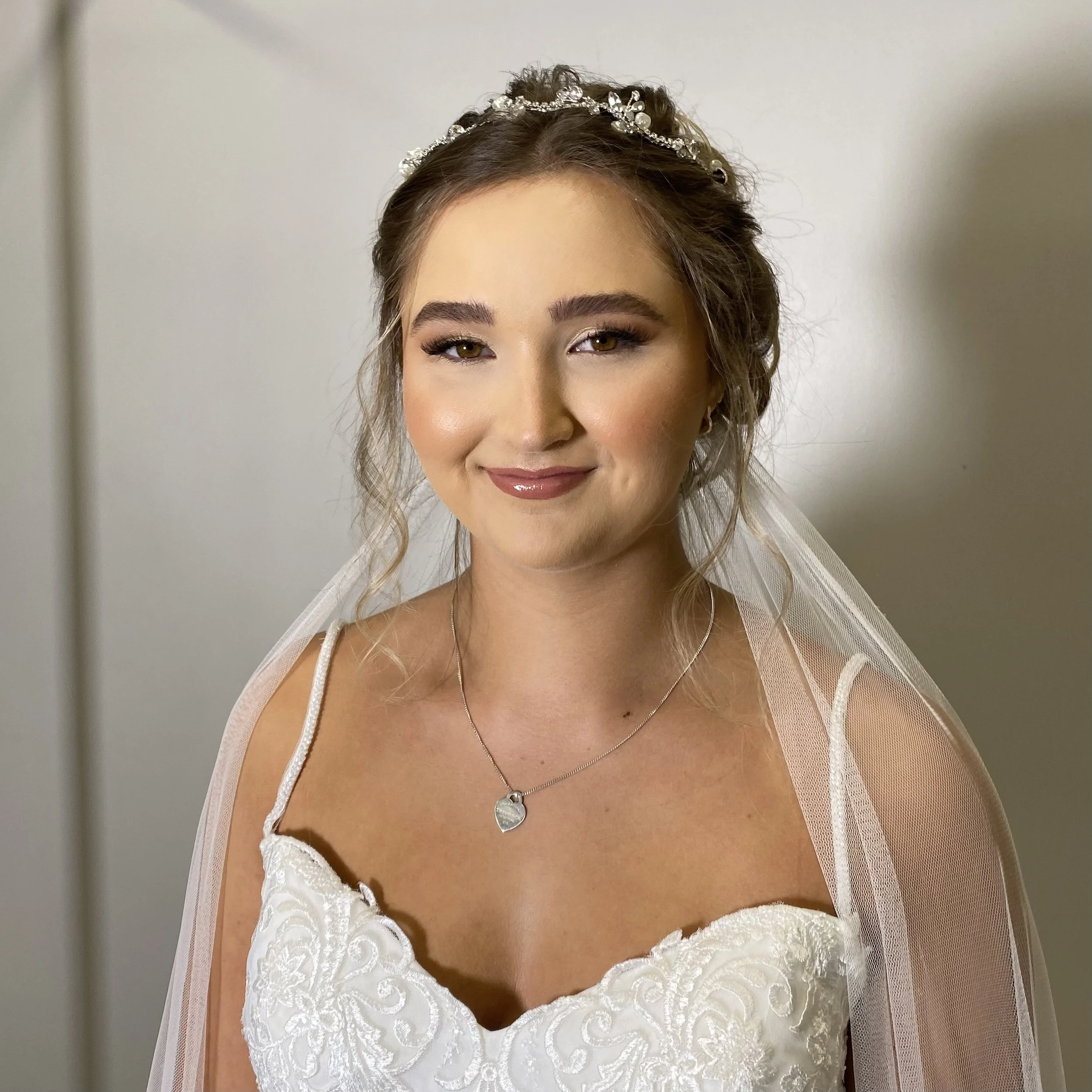 A woman with styled hair wears a tiara, white lace dress, and a delicate necklace, smiling with makeup and a sheer veil over her shoulders.