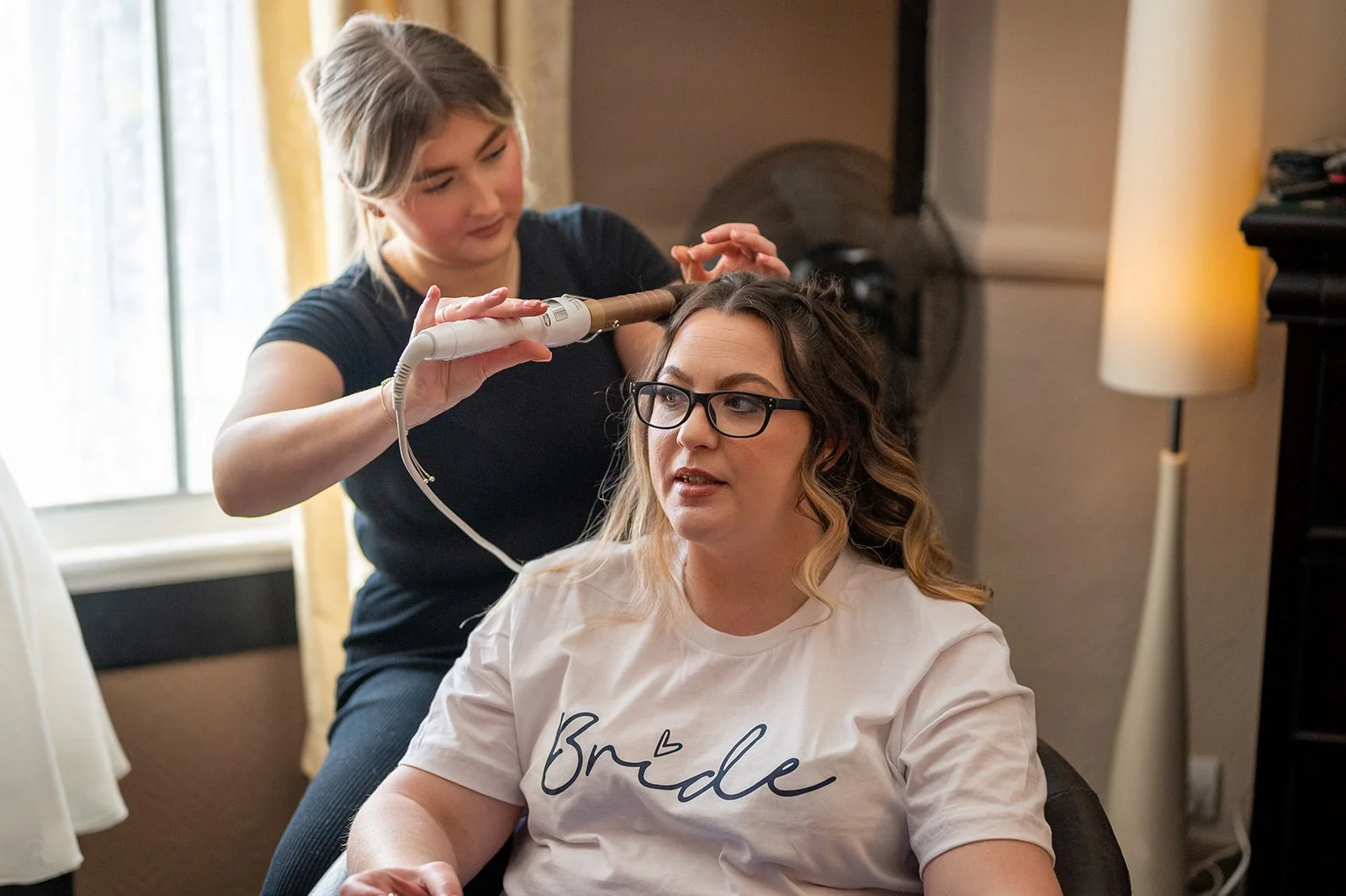 A woman with glasses sitting in a chair having her hair done by a stylist with a curling iron.