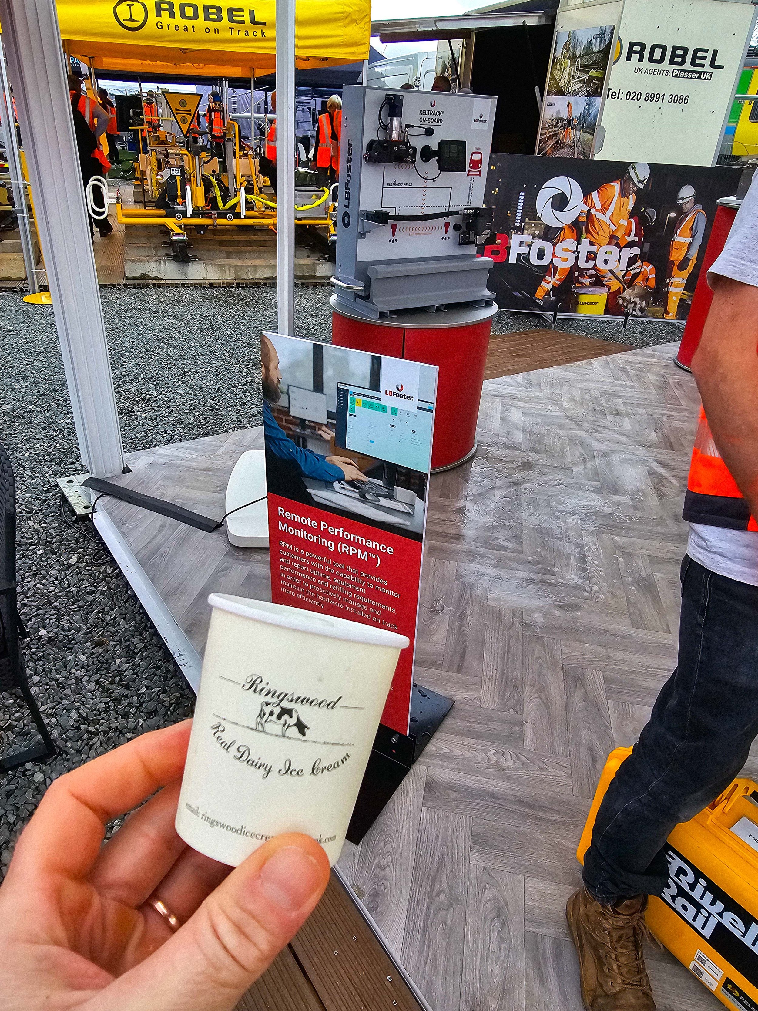 A person holding a small cup of dairy ice cream at an outdoor event or exhibition. The cup is labeled 'Ringswood Dairy Ice Cream' and features a cow logo. In the background, there are display booths, a sign for remote performance monitoring, and people in safety vests at what appears to be a construction or industrial equipment exhibition.
