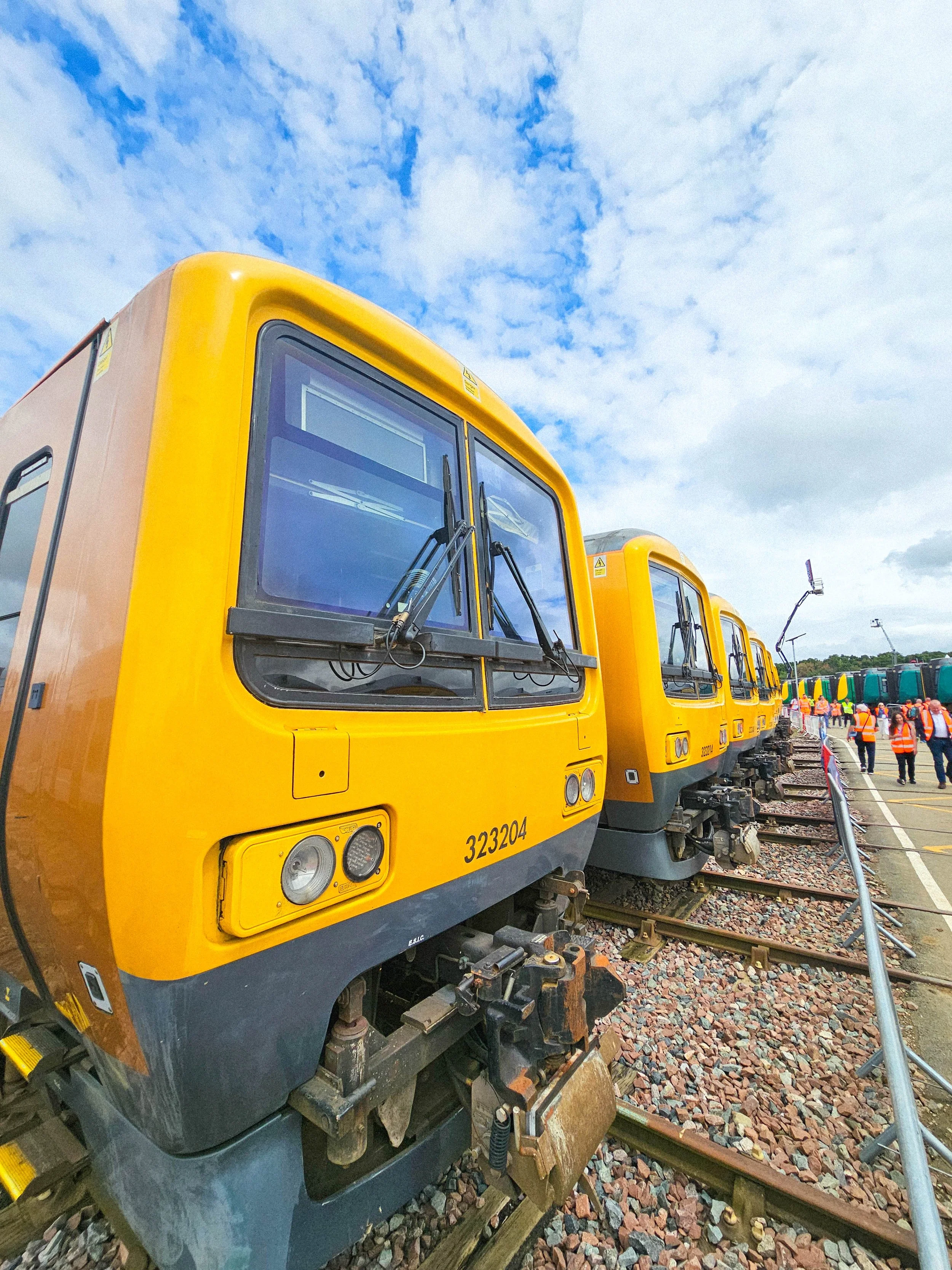 Several yellow trains parked side by side on a railway track under a partly cloudy blue sky, with people in orange safety vests walking nearby.