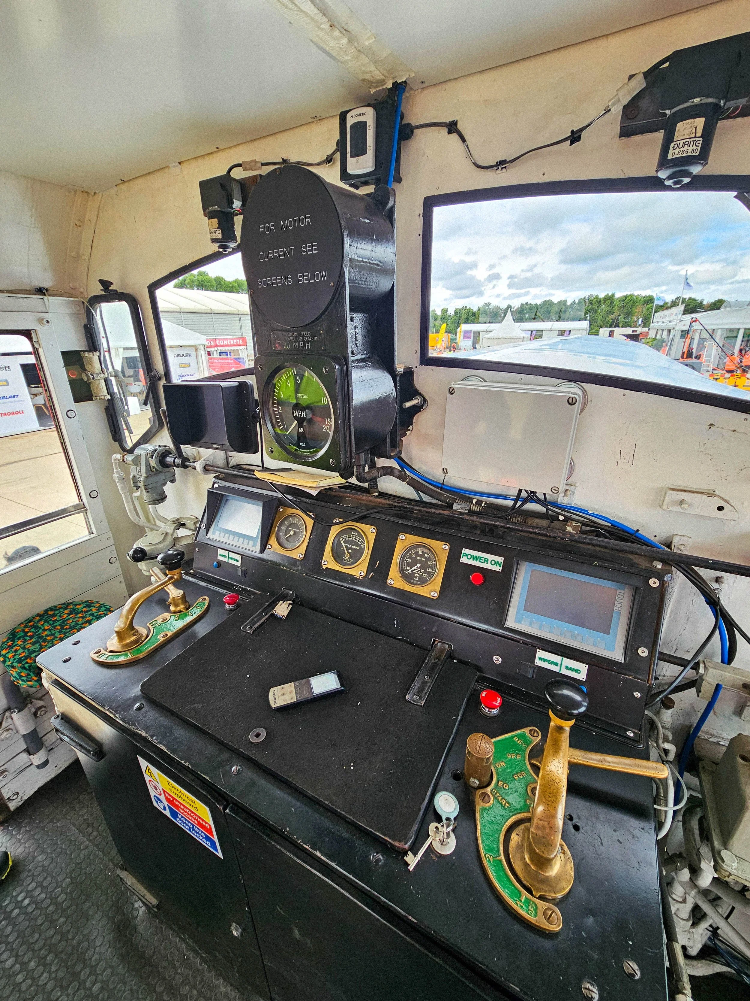 The image shows the control panel of a train cockpit with various gauges, switches, and screens, along with a view of a construction site outside the window.