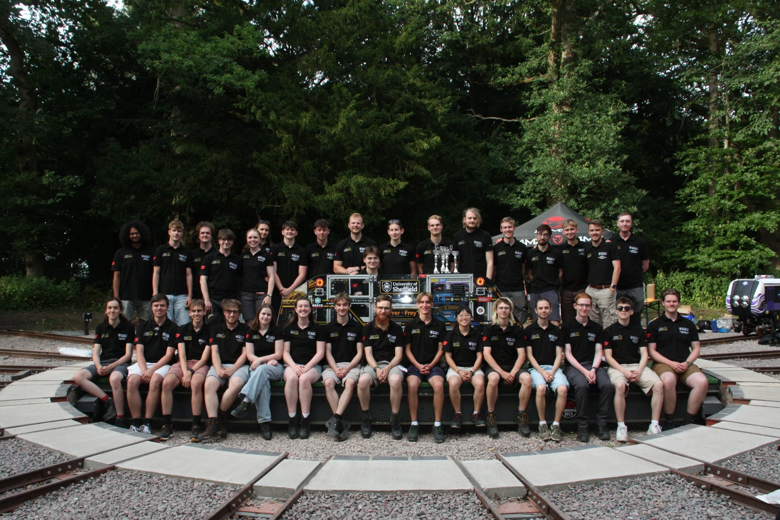 Group of young people posing outdoors for a photo, some wearing matching black shirts with logos, in front of a backdrop of trees and a tent, with some technical equipment and train tracks visible in the foreground.