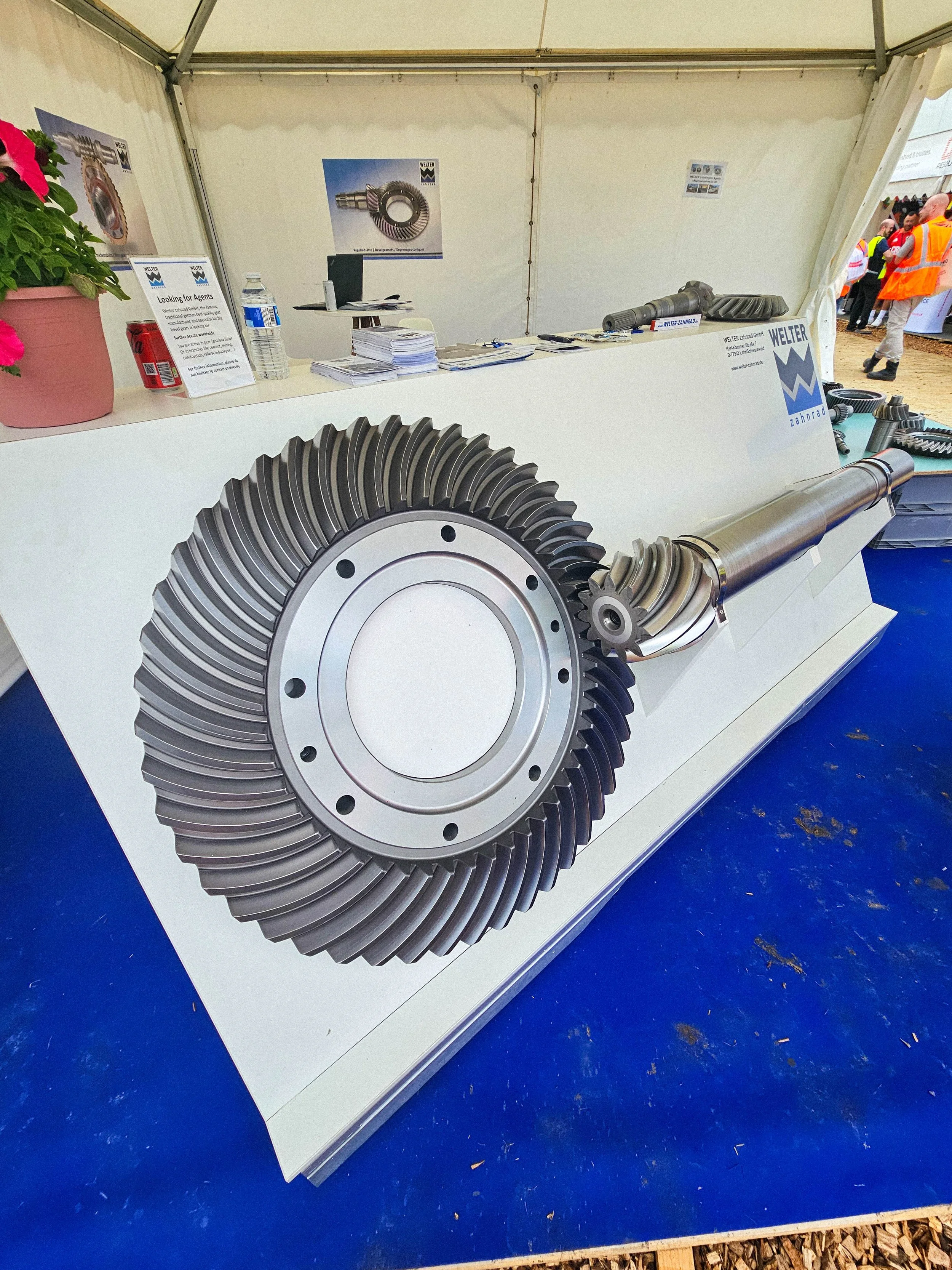 Exhibition booth displaying large, metallic gear and mechanical parts, with informational posters and printed materials on the table.
