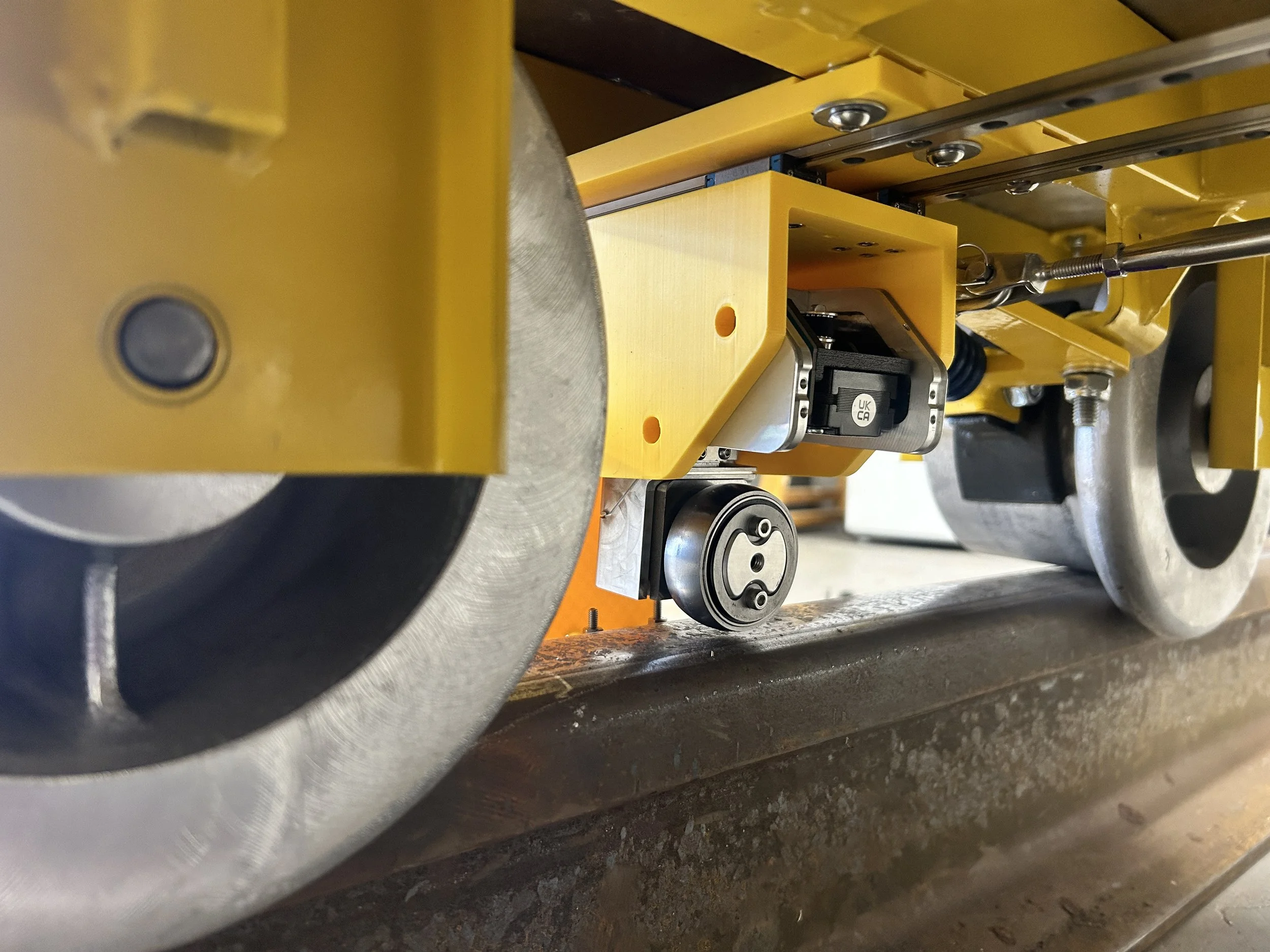 Close-up of a yellow mechanical device with metal components, including a small motor with a label marked 'UK CA,' attached to a larger metal wheel and connected with various bolts and rods.