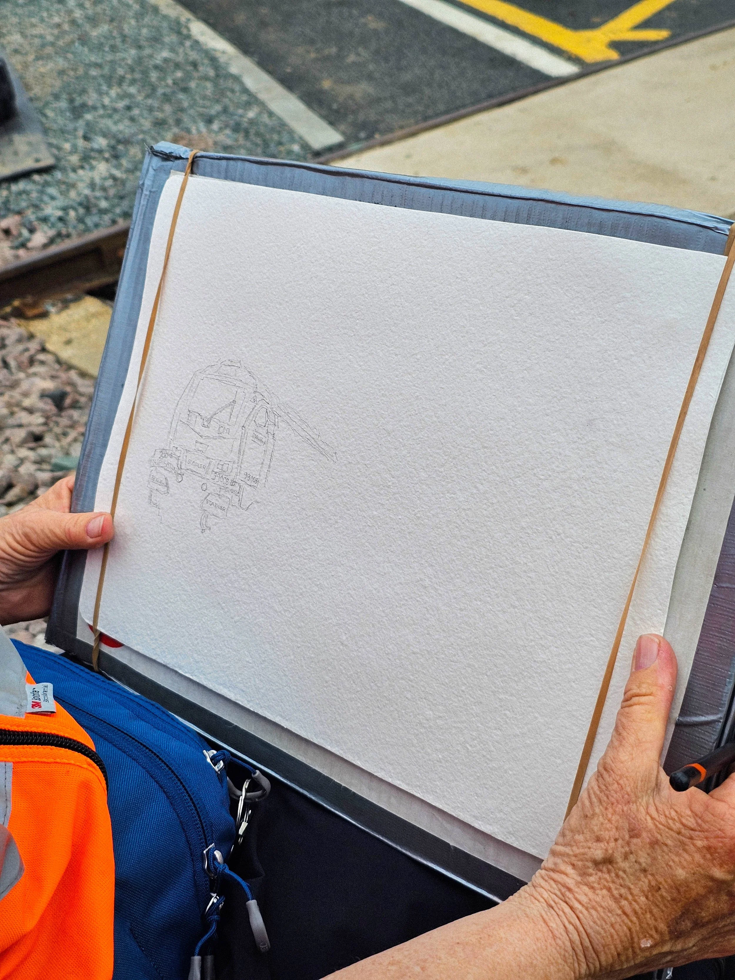 The image shows a person holding a white sketchbook or notebook with a pencil sketch of a trolleybus or tram on the page. The person's left hand is holding the book, and their right hand is visible but not holding anything. The person is wearing a bright orang navy blue backpack, and the background shows a mix of concrete and asphalt surface with painted yellow lines