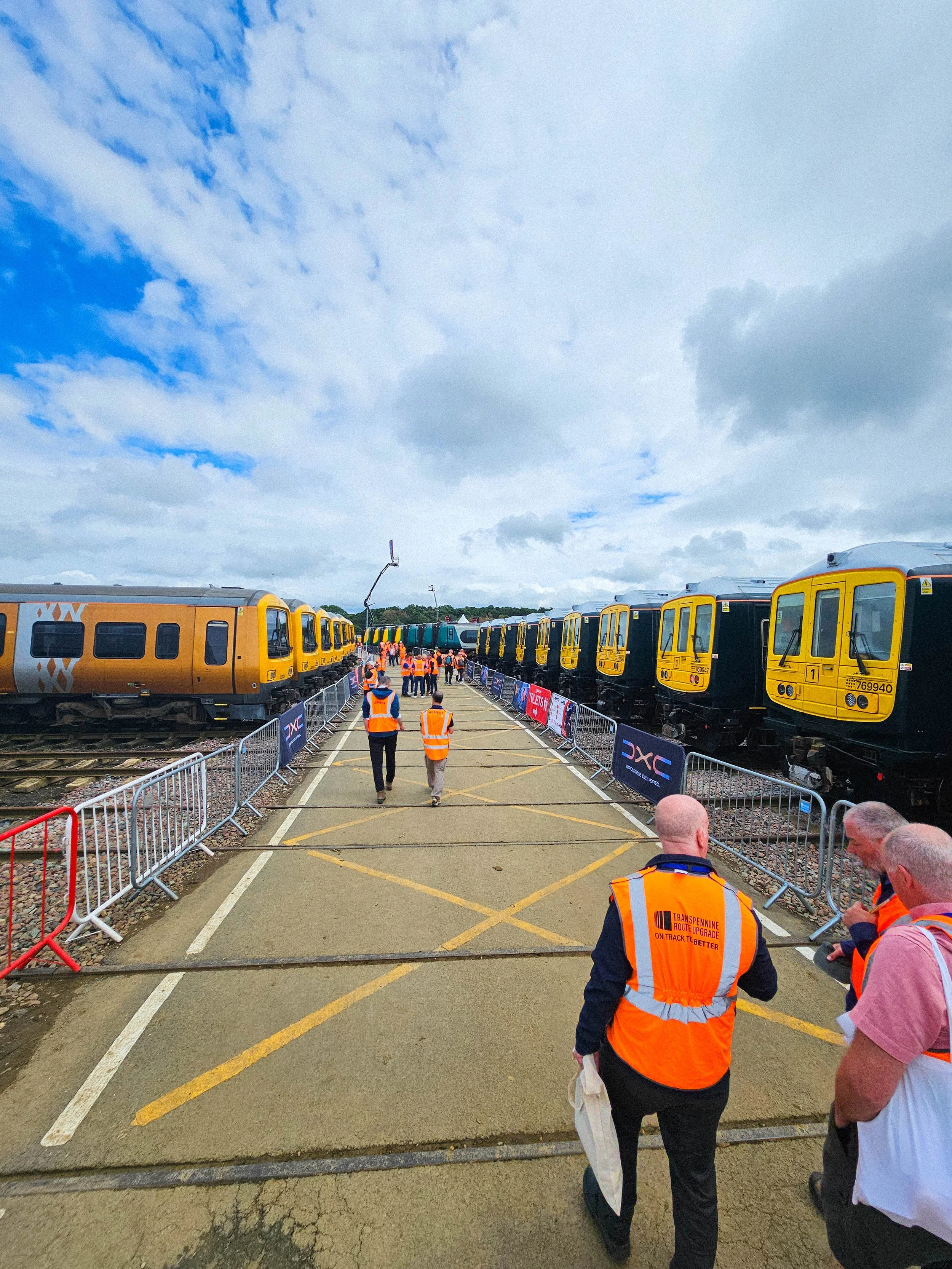 A group of people in orange safety vests walking down a train platform with yellow and black train cars on either side, surrounded by metal barriers, under a partly cloudy sky.