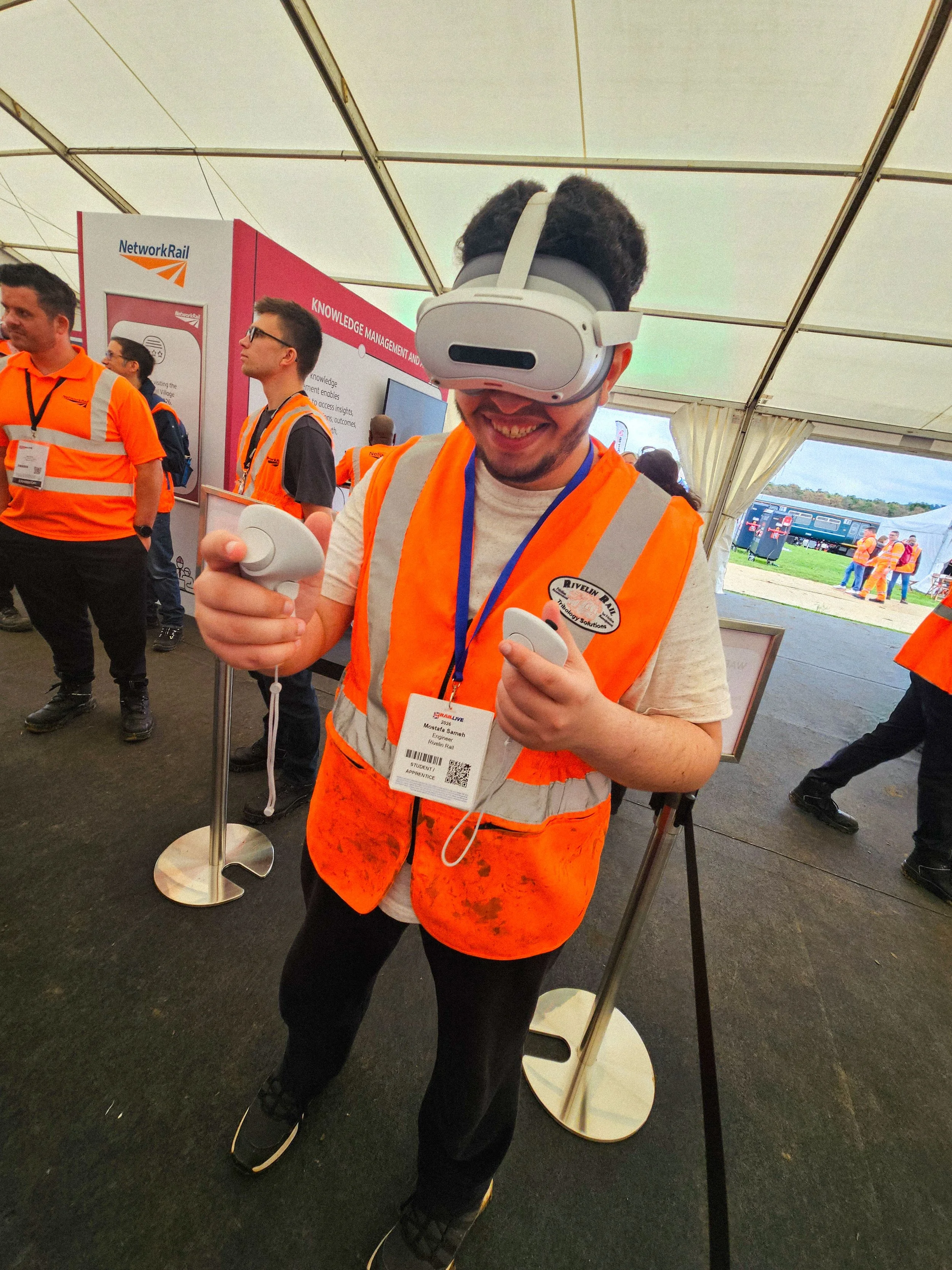 A man with curly hair and facial hair wearing an orange safety vest, a lanyard with an ID badge, and a virtual reality headset, holding VR controllers, inside a large tent at an event.