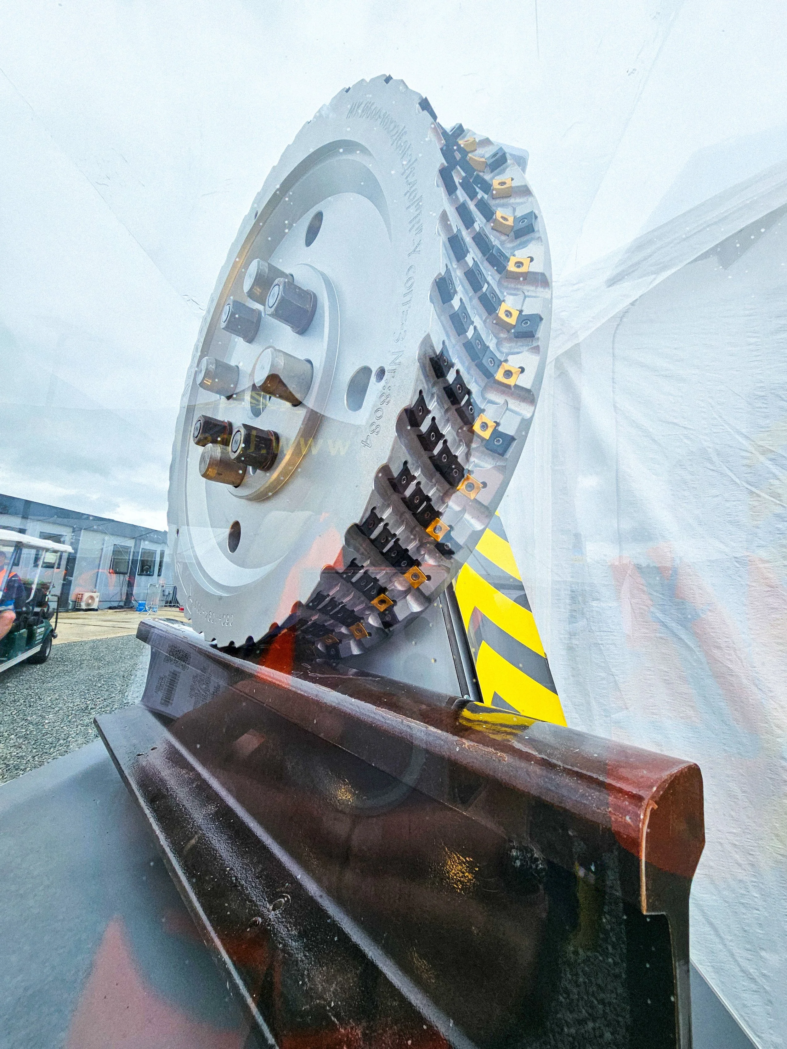 A large industrial circular saw blade with carbide-tipped teeth mounted on a machine, displayed behind a glass panel at an industrial site.