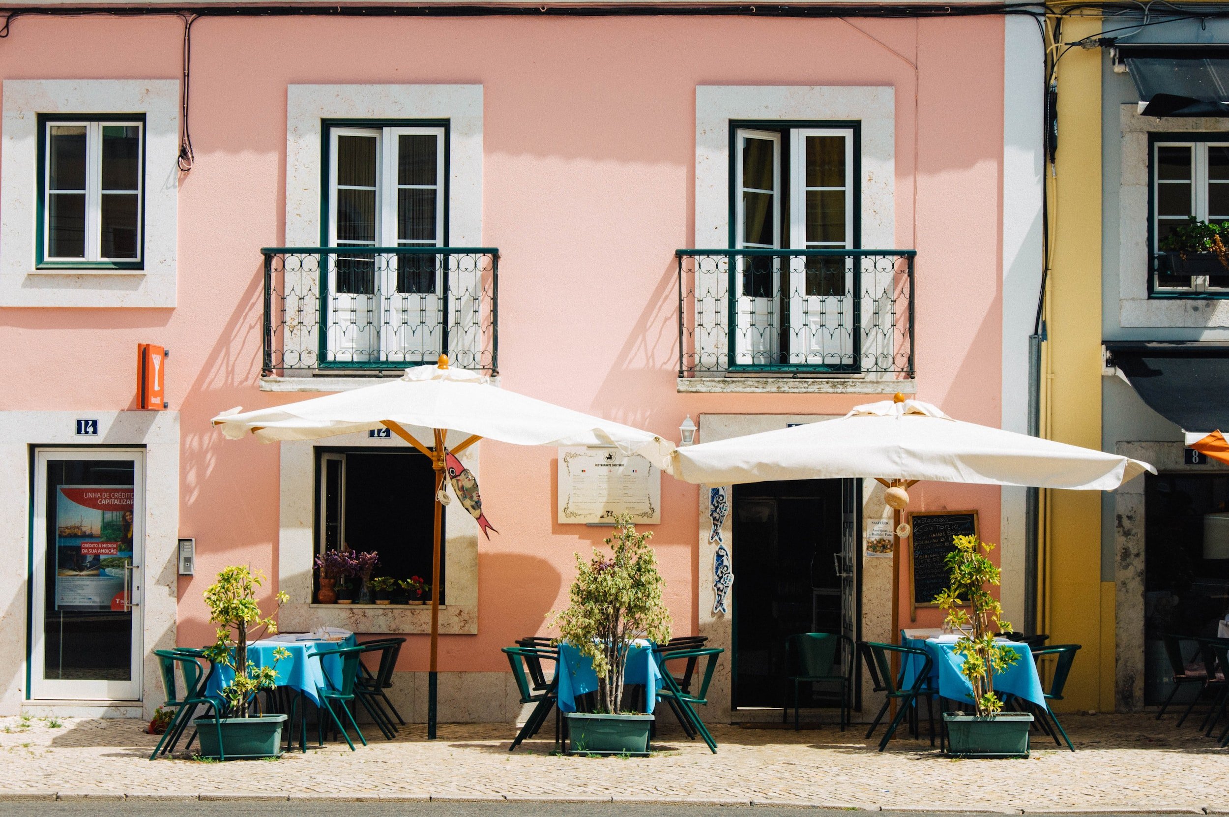 Outdoor seating area of a café or restaurant with blue tablecloths, potted plants, white umbrellas, and a pink building with two second-floor balconies in the background.
