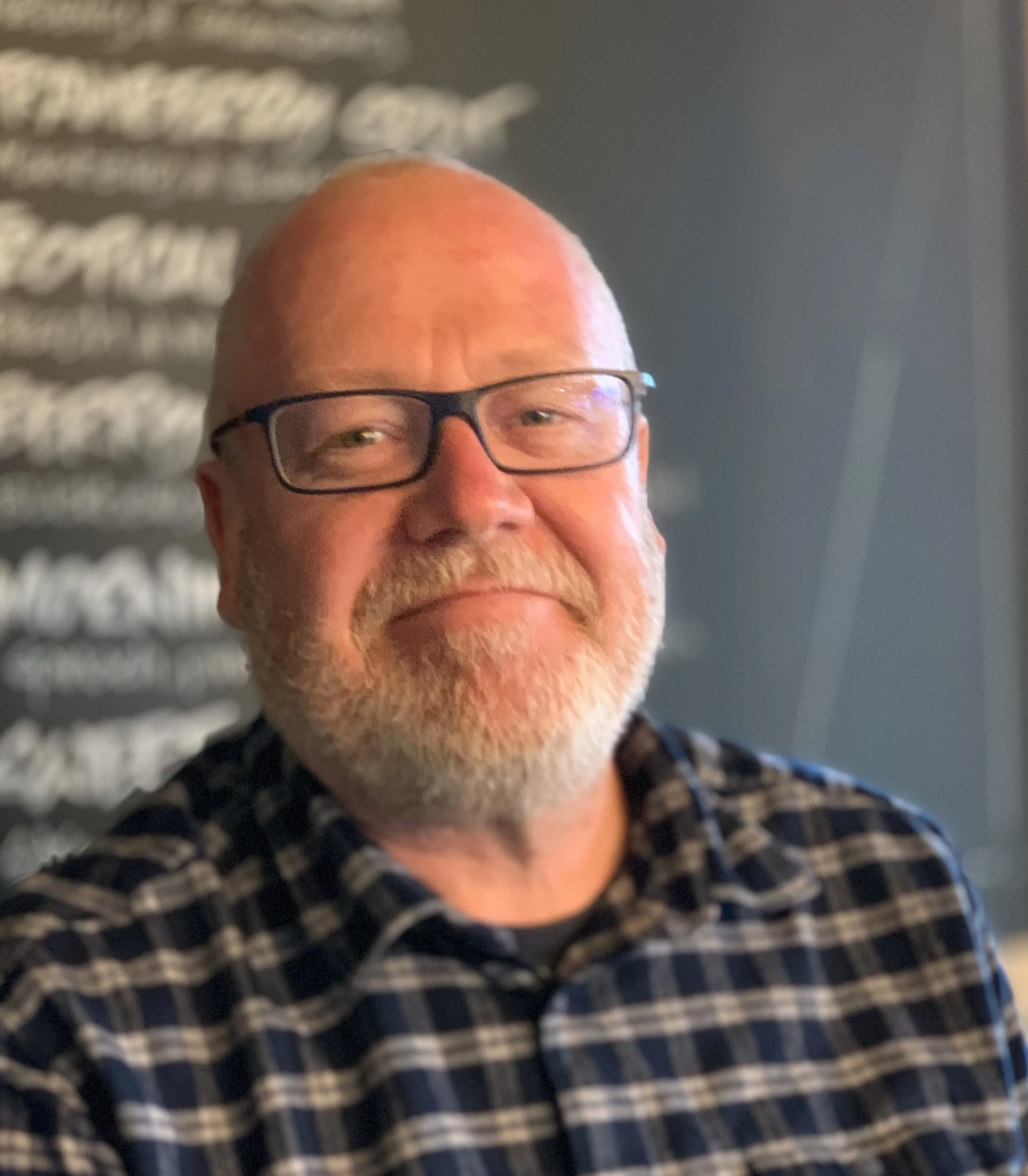 Close-up of a smiling man with glasses and a beard, wearing a checked shirt, in front of a dark grey background.