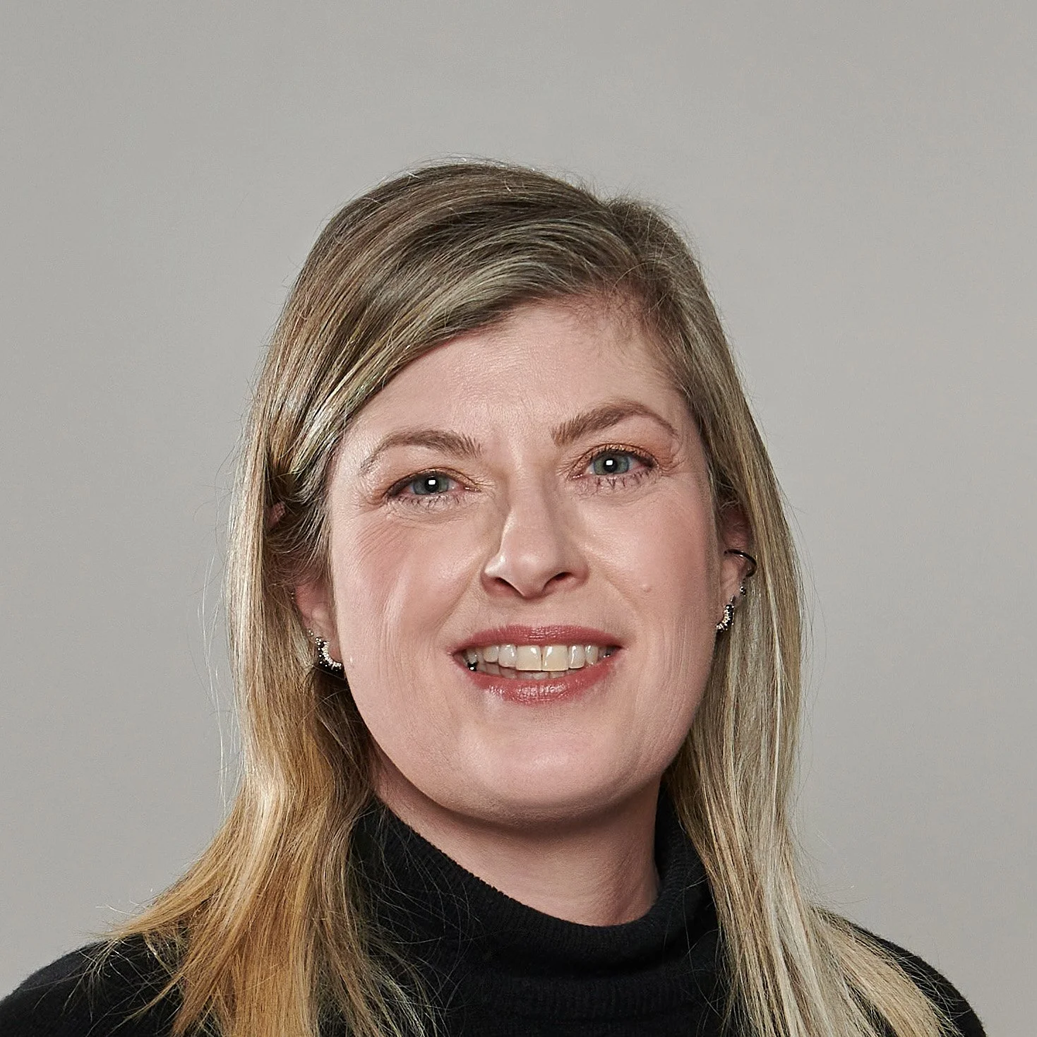 Close-up portrait of a woman with blonde hair, blue eyes, wearing small earrings and a black turtleneck, smiling against a plain grey background.