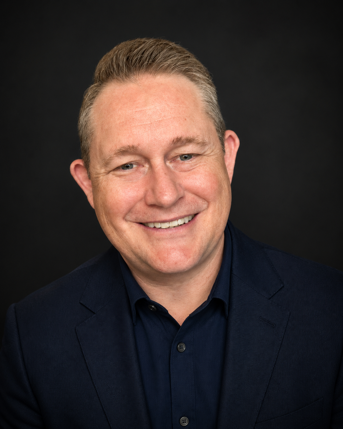 Headshot of a smiling middle-aged man with short blond hair, wearing a dark blue suit and a black shirt, against a black background.