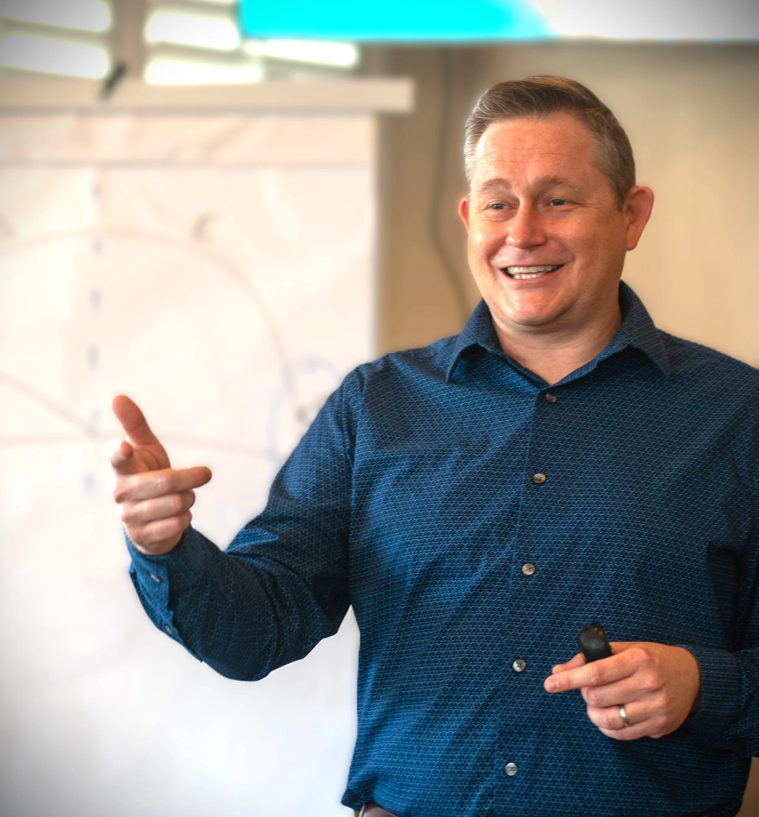 A man wearing a dark blue, patterned button-up shirt holds a marker while smiling. His right hand is giving a thumbs-up gesture, and he is positioned in front of a whiteboard or screen with a blurred background.