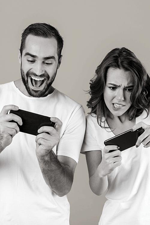Black and white image of two people in white shirts playing mobile games. The man looks excited, while the woman appears frustrated and intense.