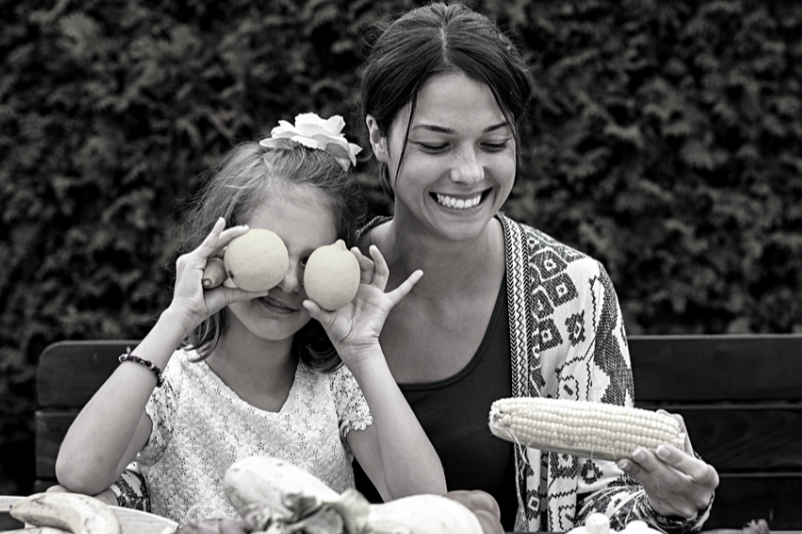 Black and white image displaying a mom and her child. The little girl is holding two lemons in front of her eyes