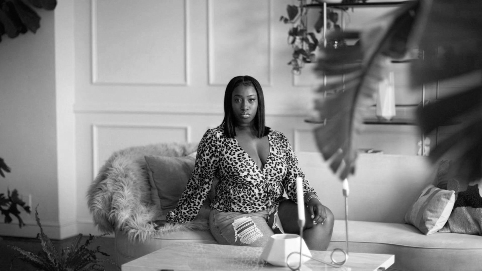 Black woman sitting on a sofa in a modern living room, wearing a leopard print top and ripped jeans, with large leaves of a plant partially visible in foreground.