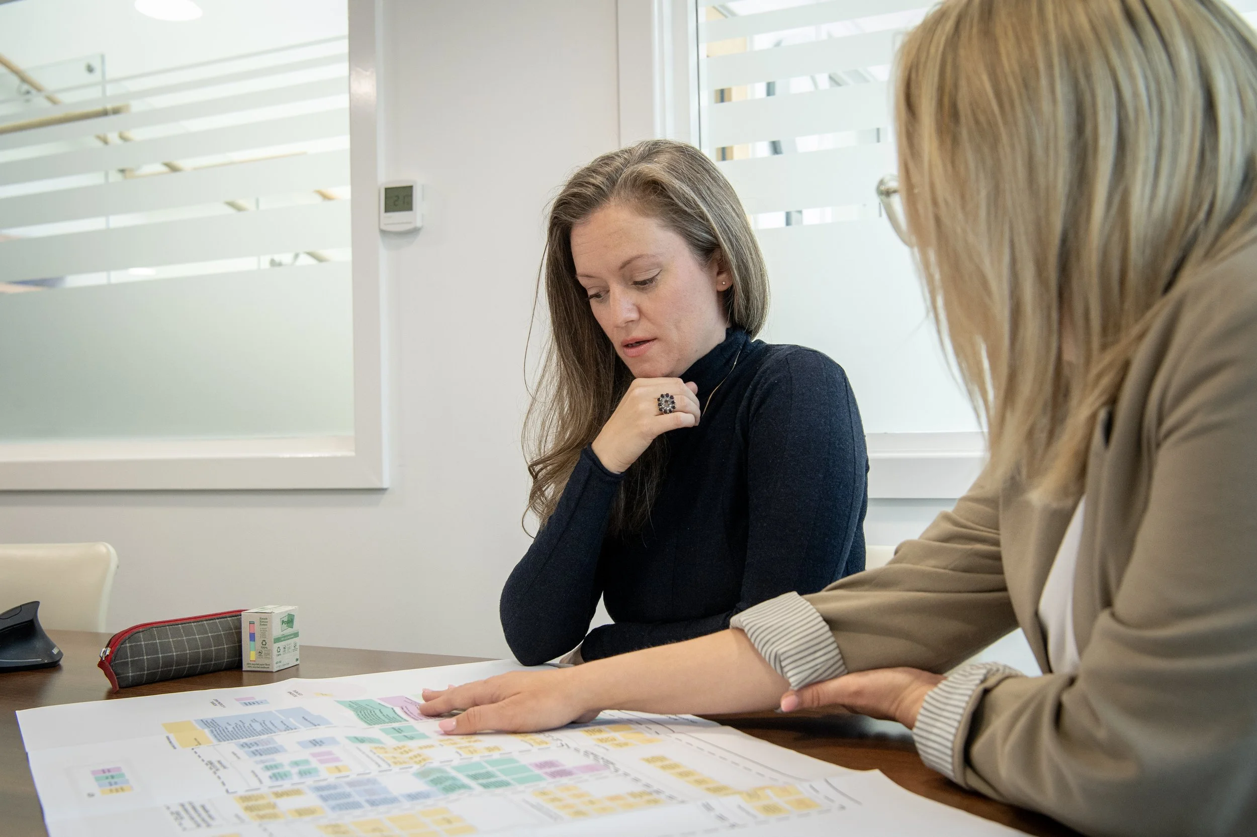 Two women sitting at a table discussing a large map or chart, with a brown pencil case, a medicine box, and a computer mouse on the table.