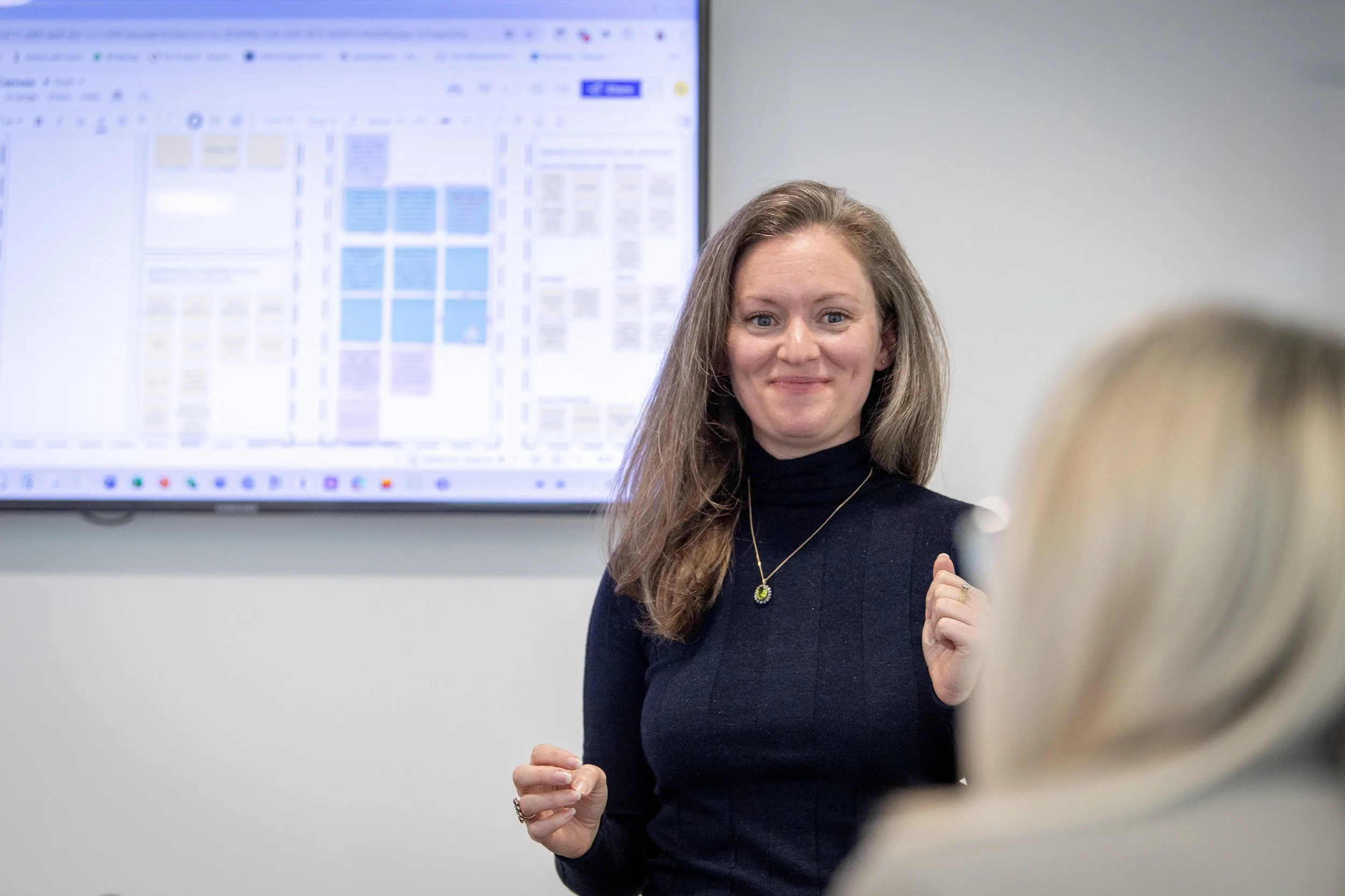 Woman with long hair in a black turtleneck and necklace standing in front of a display screen, smiling during a presentation or meeting.
