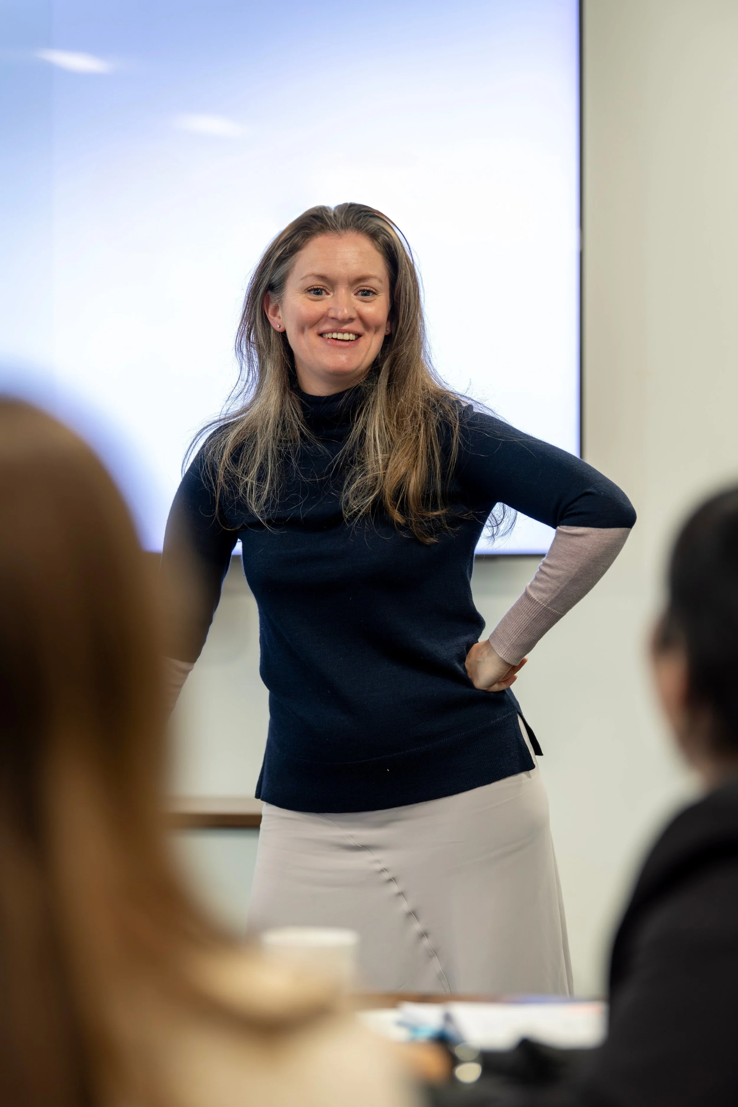 A woman with long brown hair, wearing a black and beige sweater, smiling and standing in front of a large screen, with an audience in the foreground.