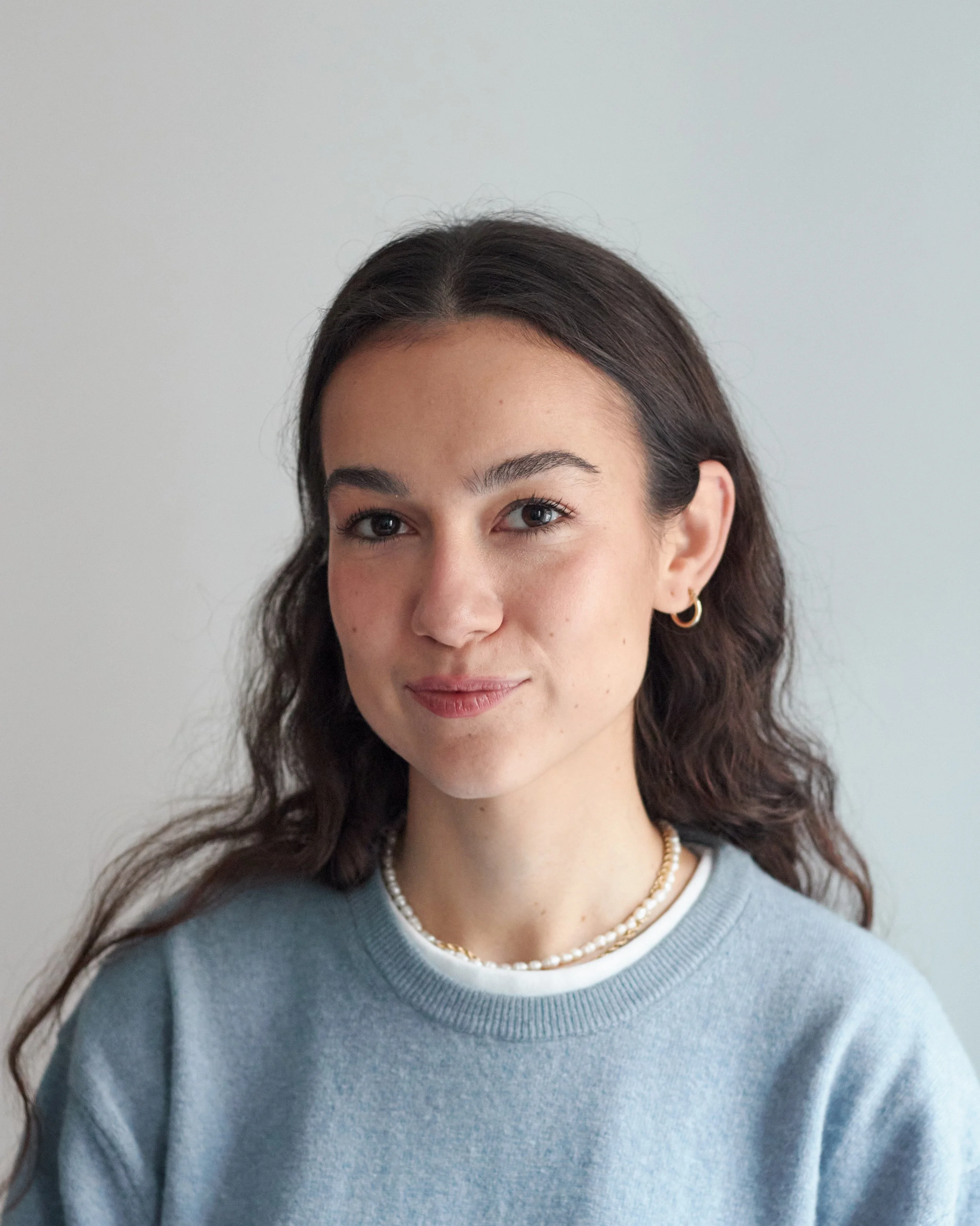 Een vrouw met lang, golvend bruin haar kijkt in de camera, voor een witte achtergrond.