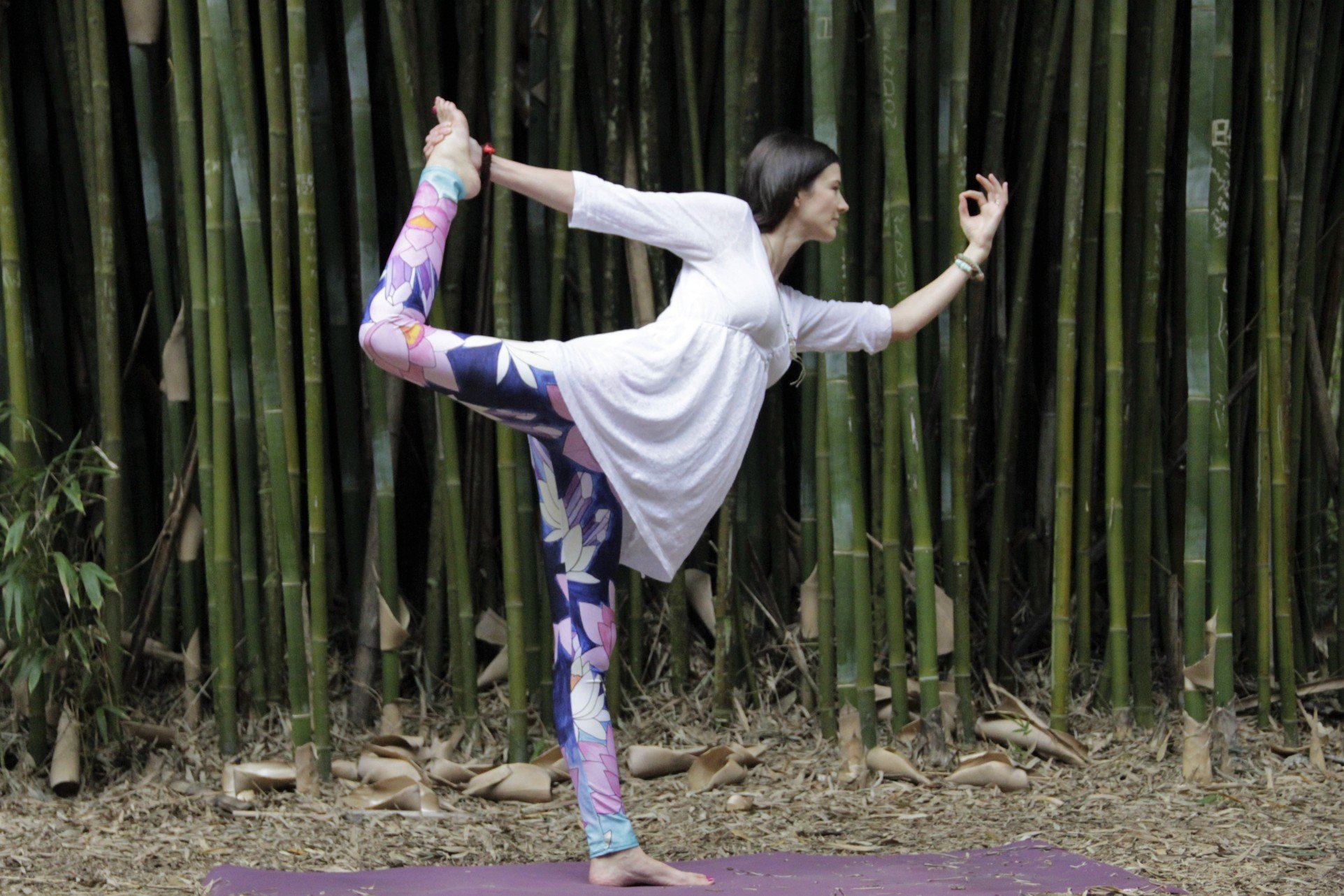 Woman wearing yoga pants and a white shirt in dancing shiva pose against a background of bamboo.