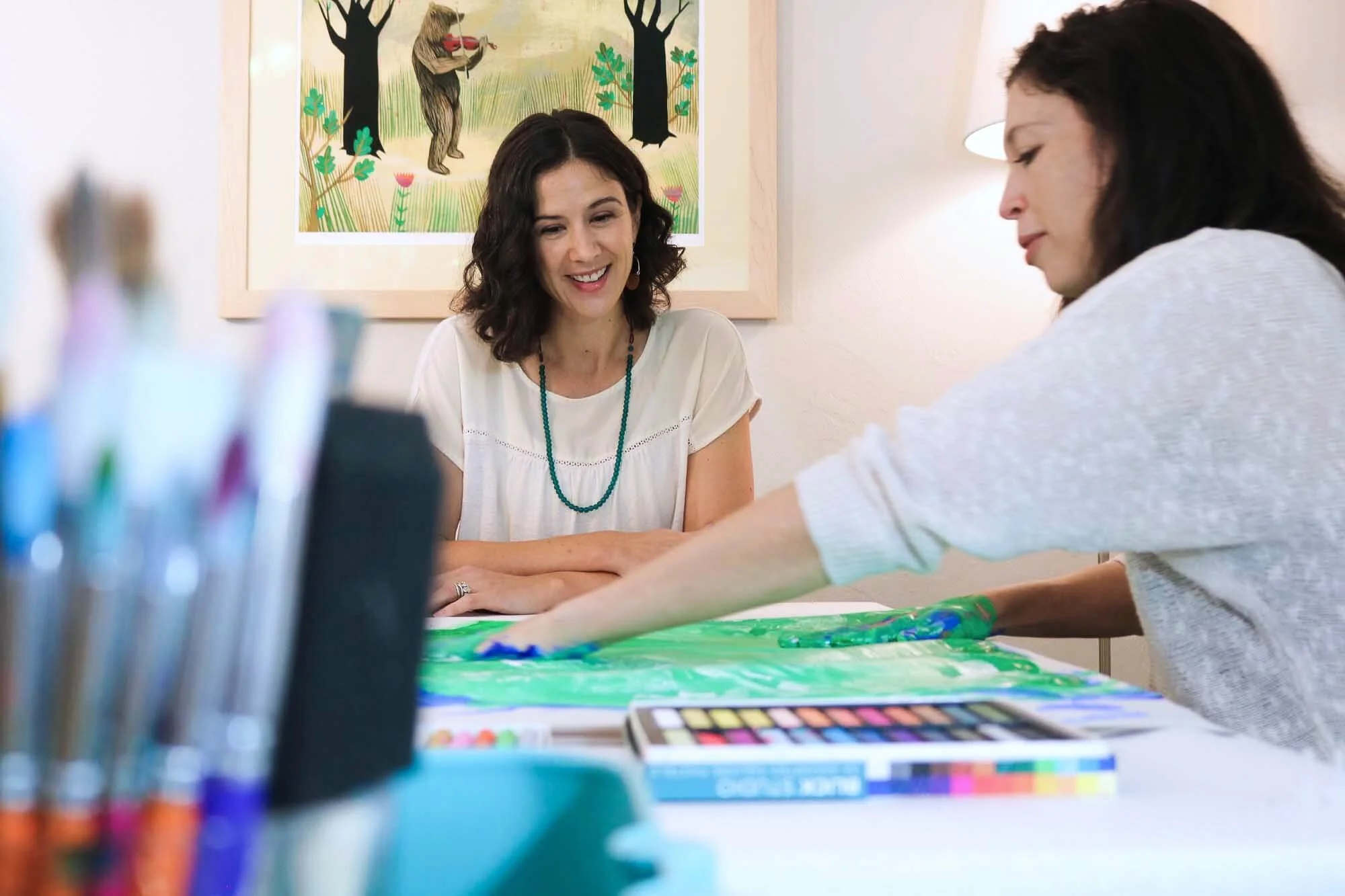 Two women are seated at a table engaging in an art activity. One woman, with curly dark hair and a white top with a turquoise necklace, smiles as she watches the other woman, who is reaching across the table. The table has paints and art supplies, and there is a colorful painting with trees and a bear in the background.
