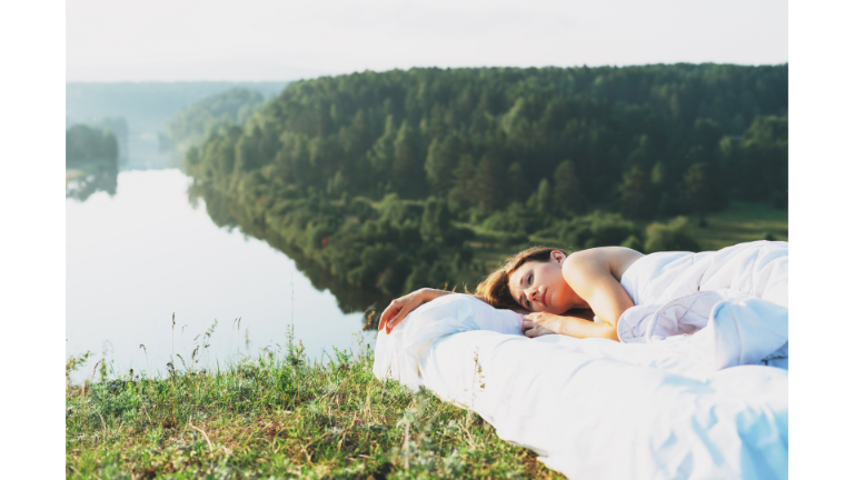 Woman lying on her side on a mattress with white sheets outside with trees and a river in the background.