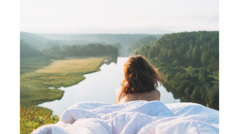 Woman lying on her stomach facing away from the viewer on a mattress with white sheets outside with trees and a river in the background.