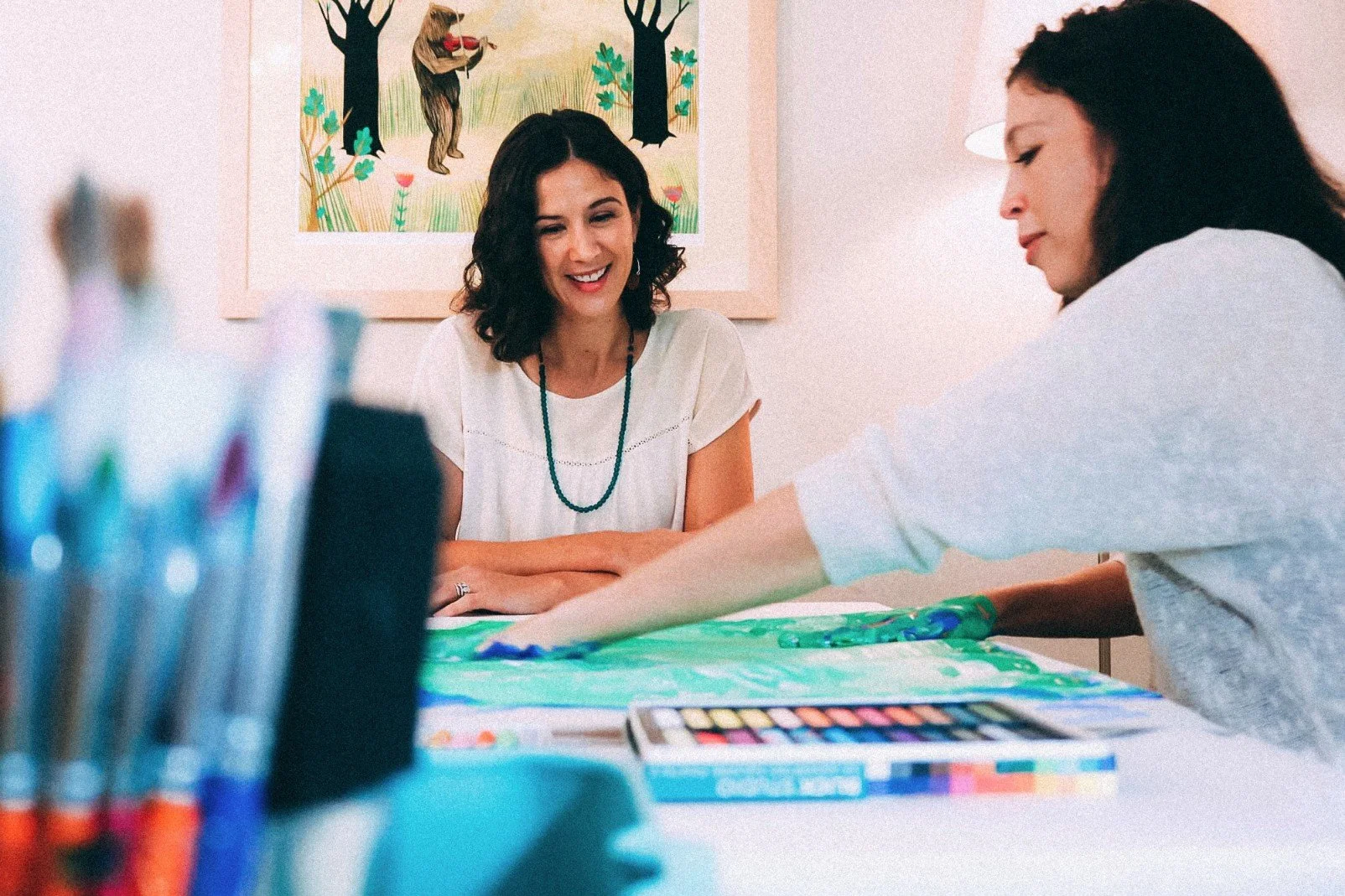 Two women seated at a table finger painting with green and blue paint..