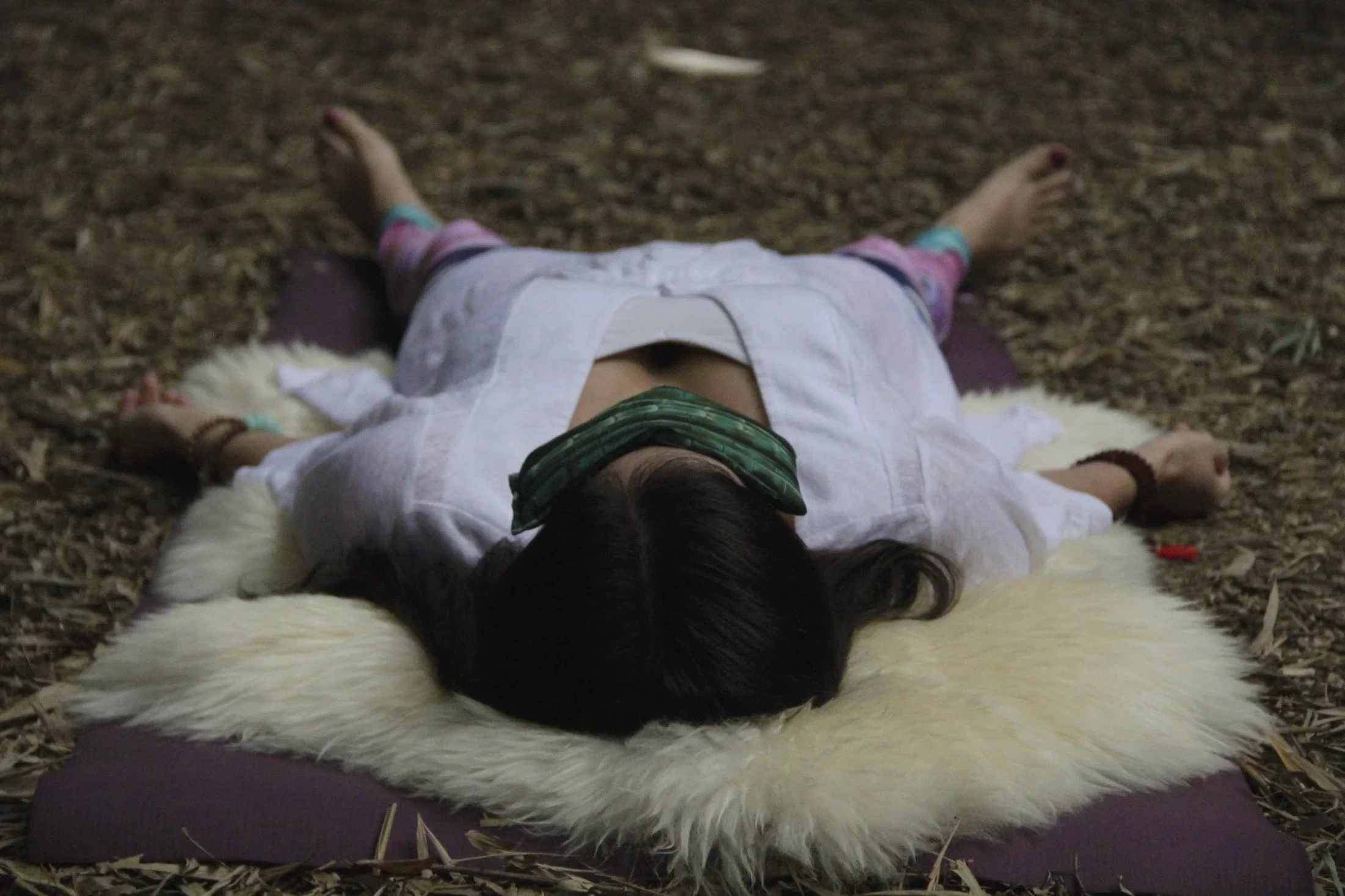 Woman lying on a sheepskin on the ground in corpse pose with an eye pillow over her eyes.