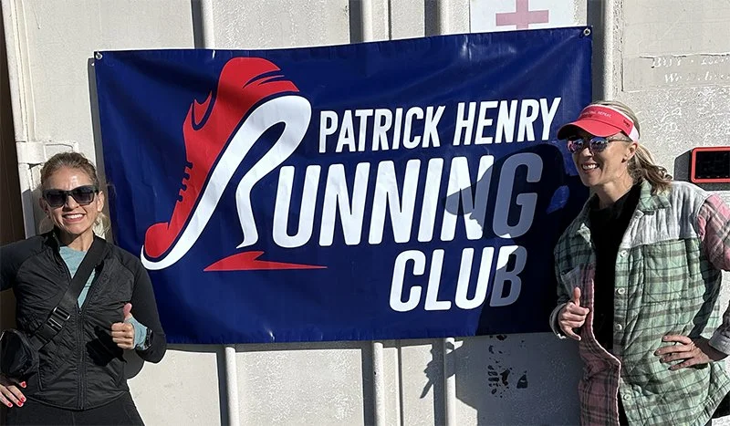 Two women standing next to a large blue banner with a running shoe graphic, reading "Patrick Henry Running Club." Both women are smiling and giving thumbs-up gestures.