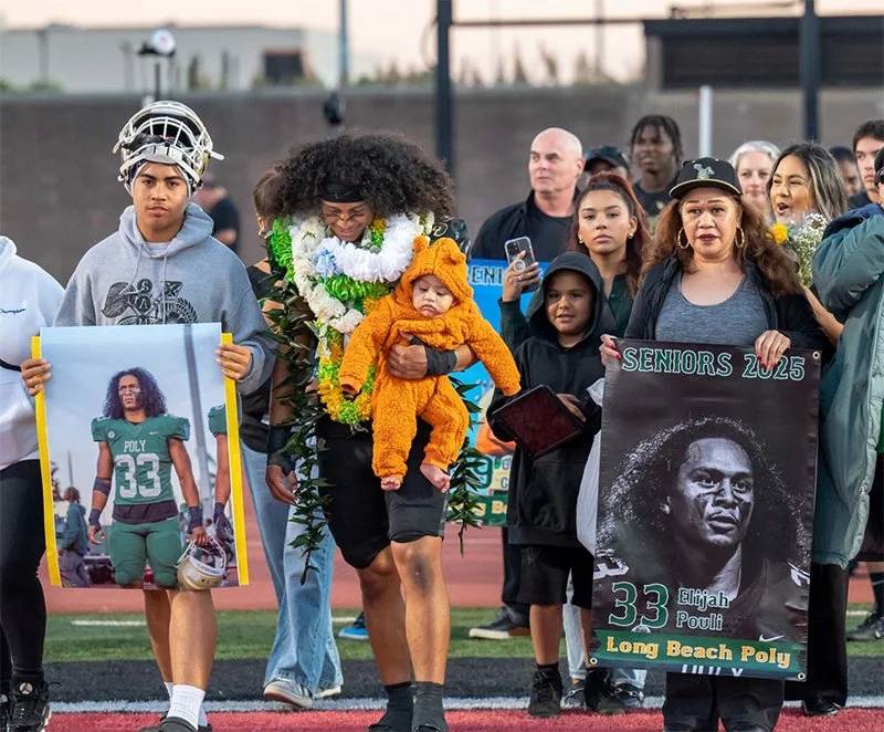 Group of people, including a woman carrying a baby in a lion onesie, participating in a memorial or graduation event at a track field. One person holds a portrait of a football player Elijah Pouli with the caption "Seniors 2025" and "Long Beach Poly." Others hold signs, and some are taking pictures.