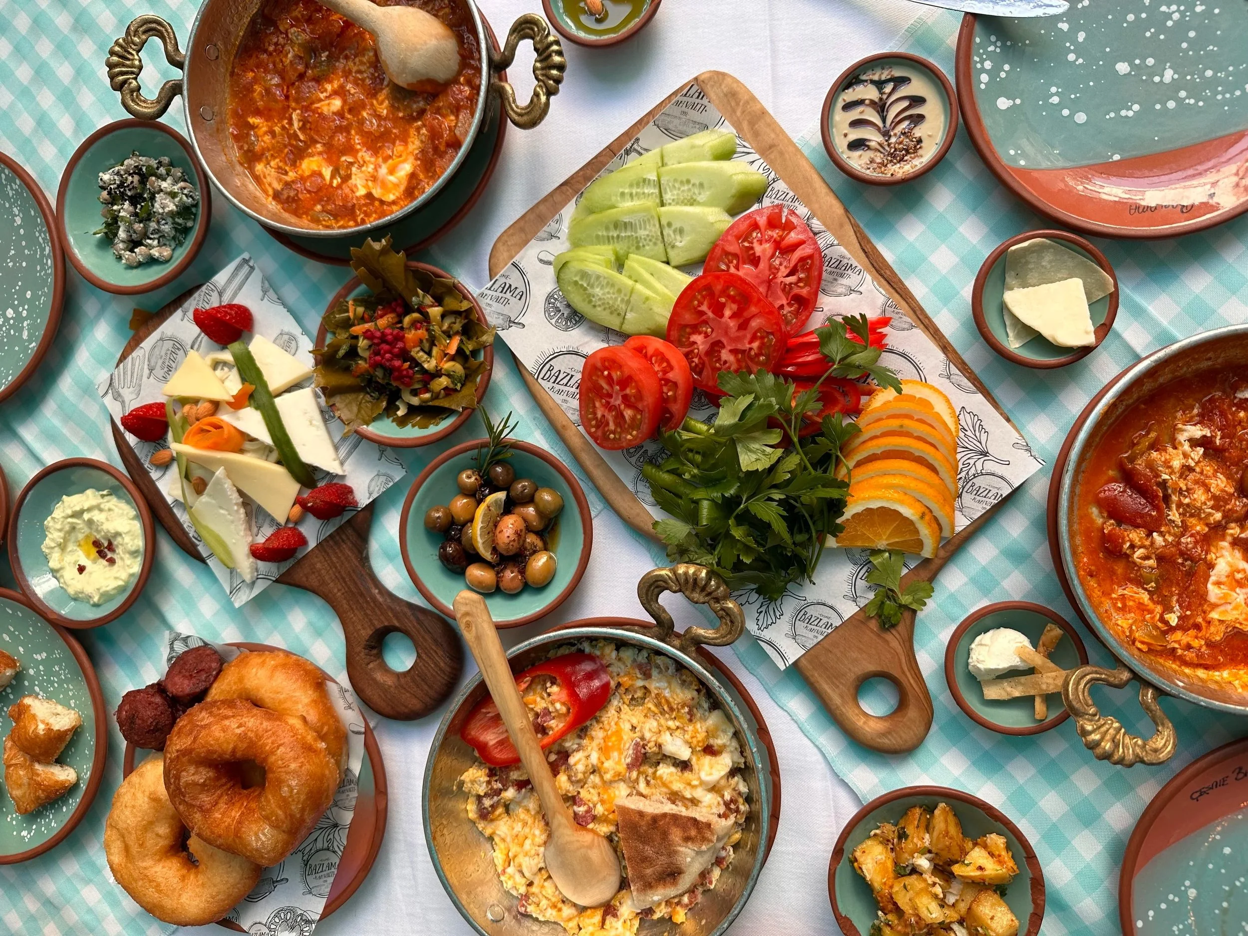 A spread of Mediterranean dishes including tomato, cucumber, and orange slices, olives, salads, dip, bread, and other small appetizers on a checkered tablecloth.