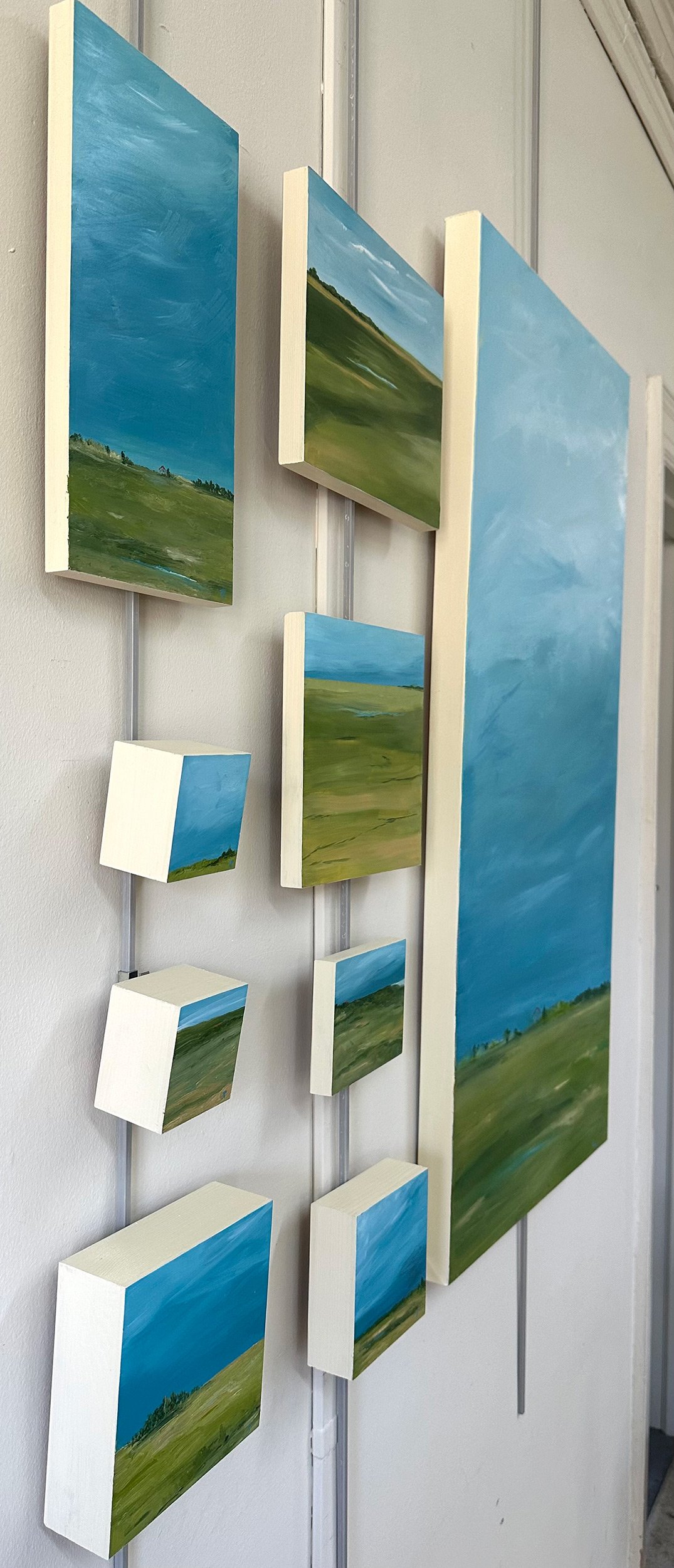 Angled, side view of a grouping of 9 paintings in a variety of sizes, hanging on a wall. They all feature turquoise skies, green grass, and have an abstracted vibe.
