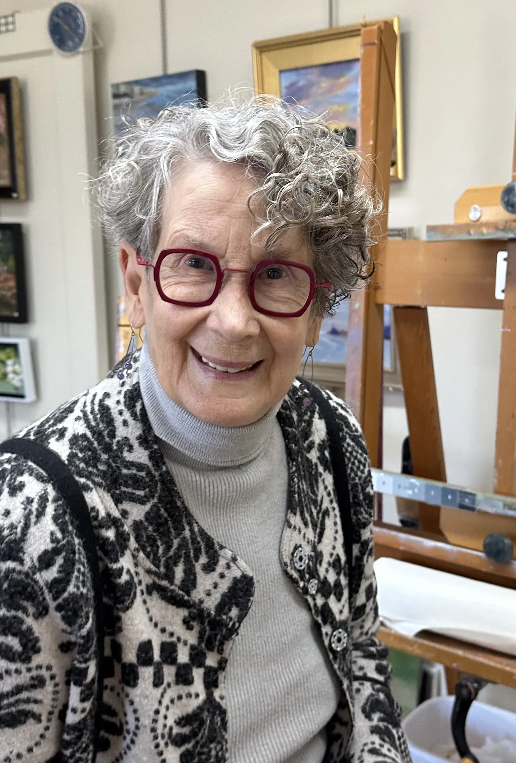Photo of smiling lady - Phyllis Sharpe - with short, curly, assymmetrically-cut hair. She's wearing dark red square glasses, gray turtleneck, and a black and white wool coat with unique pattern.  She's in her fine art studio with easel behind her.