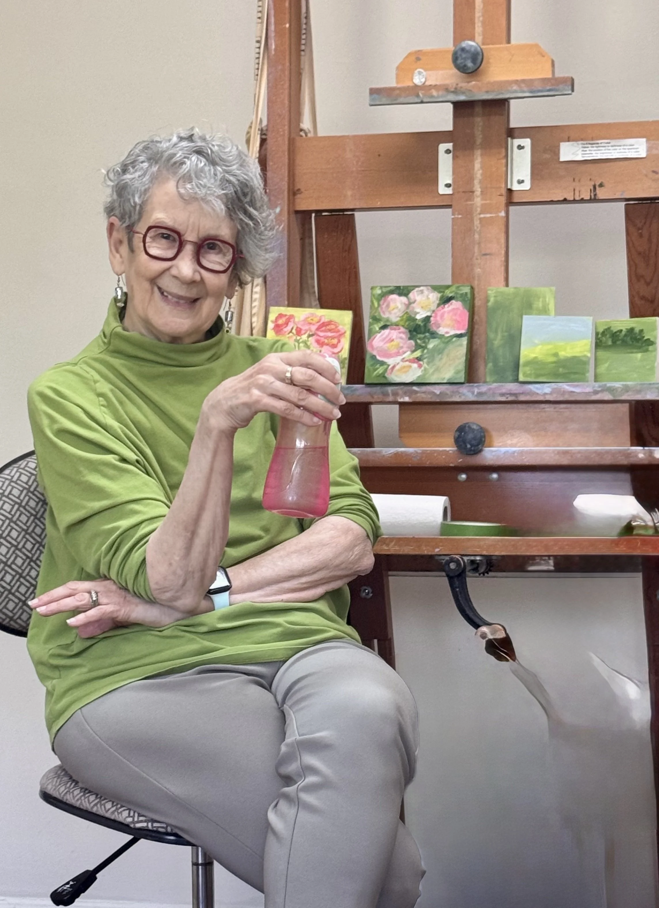 Photo of a smiling lady with short curly, asymmetrically-cut hair. She's wearing artsy squarish dark red glasses and a lime green turtleneck shirt. She's sitting at an easel where a display of small paintings have been displayed.