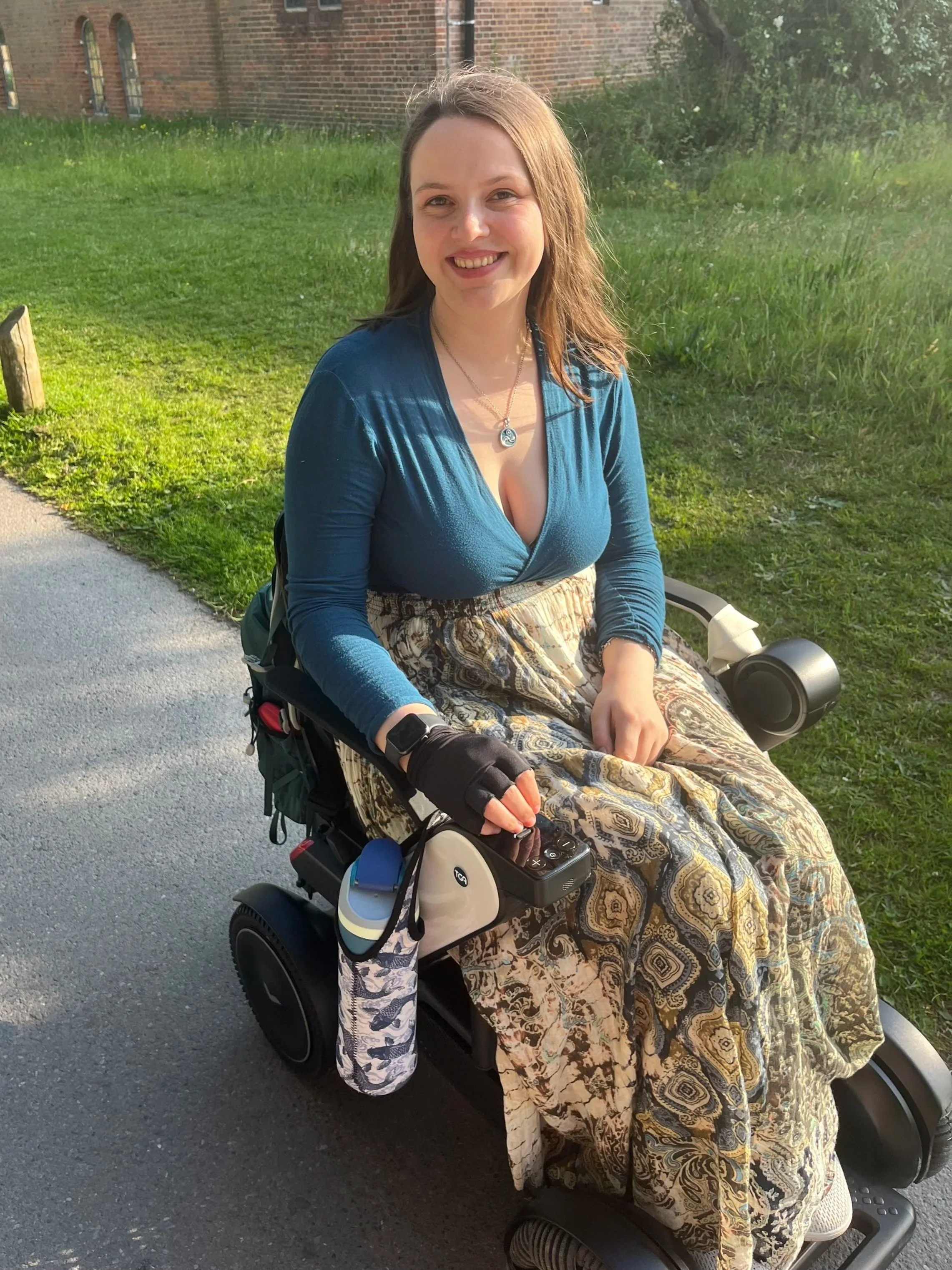 Blossom, a brunette white woman, sitting in a wheelchair outdoors on a sunny day, smiling at the camera. She is wearing a blue top, patterned skirt, and a necklace. Behind her is a grassy area and a brick building.