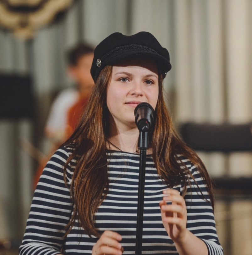 Blossom, a brunette white woman, wearing a navy and white striped shirt and a black hat, standing in front of a microphone during a speech, with a blurred stage curtain in the background.