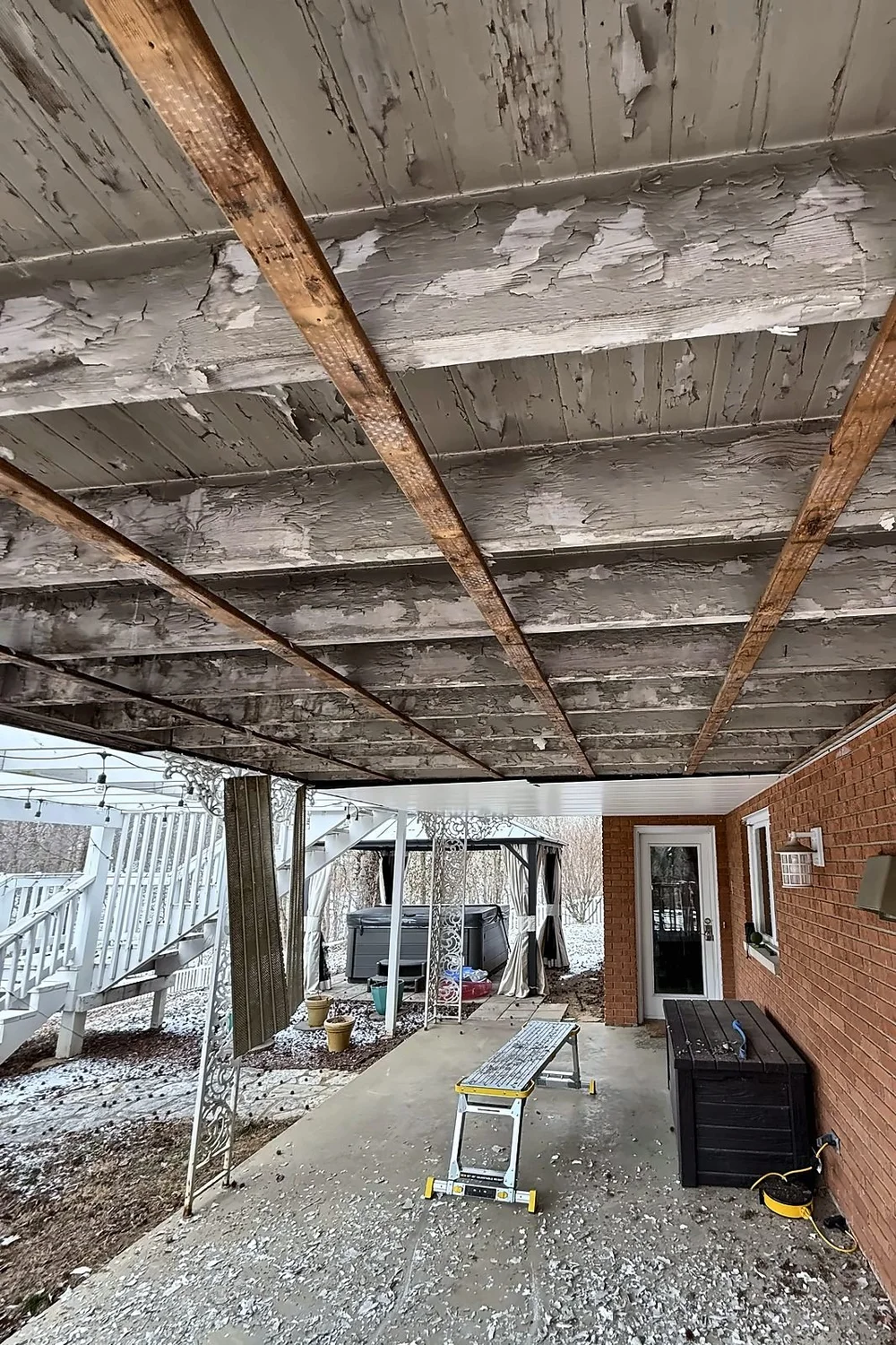 Underside of an upper deck after the old soffit was removed, showing bare joists with peeling paint over a basement patio