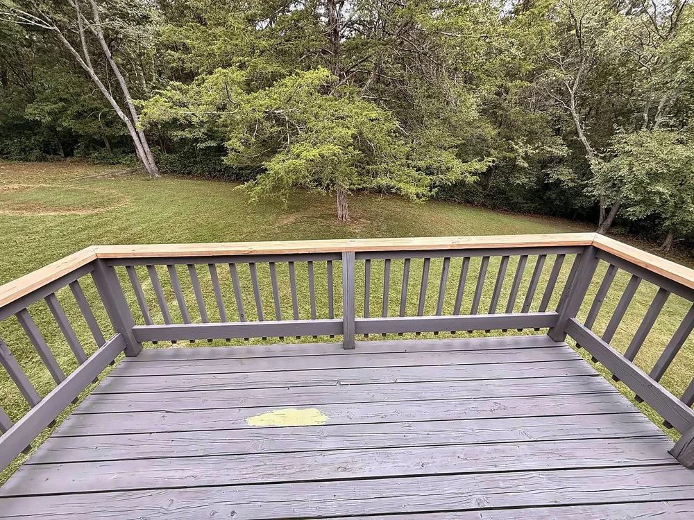 Gray deck railing with new pressure-treated top caps, looking out over a wooded backyard