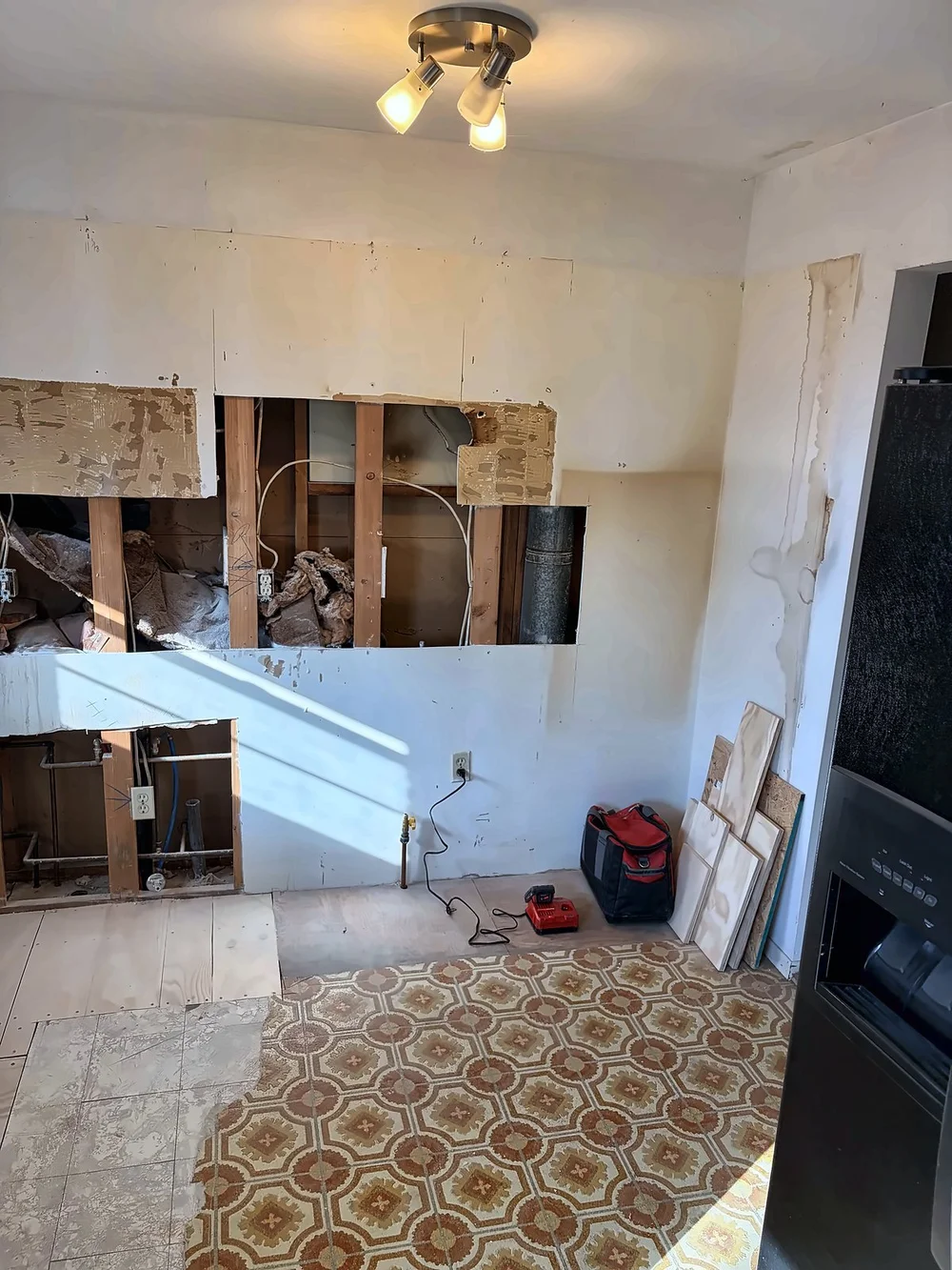 Gutted kitchen with the drywall cut out along the counter wall for the backsplash base, old patterned vinyl flooring and a black refrigerator