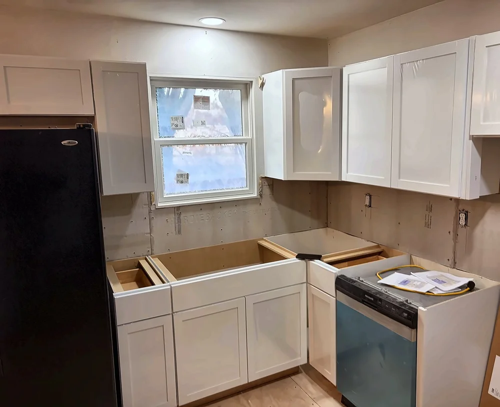 New white shaker base and wall cabinets being installed around the kitchen window with a new dishwasher in place