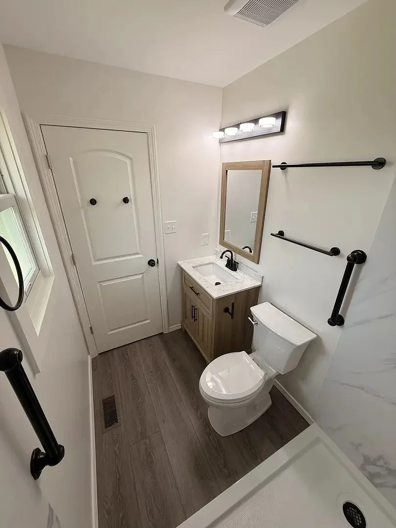 Modern bathroom with a toilet, wooden vanity, mirror, and lighting. White walls, wood floor
