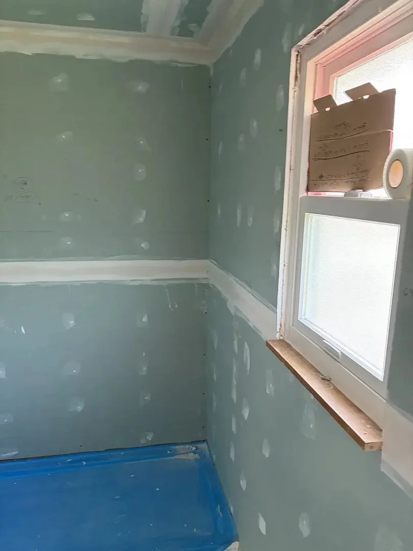 A room under construction with drywall, window, and blue floor covering. Cardboard and tape are on the window