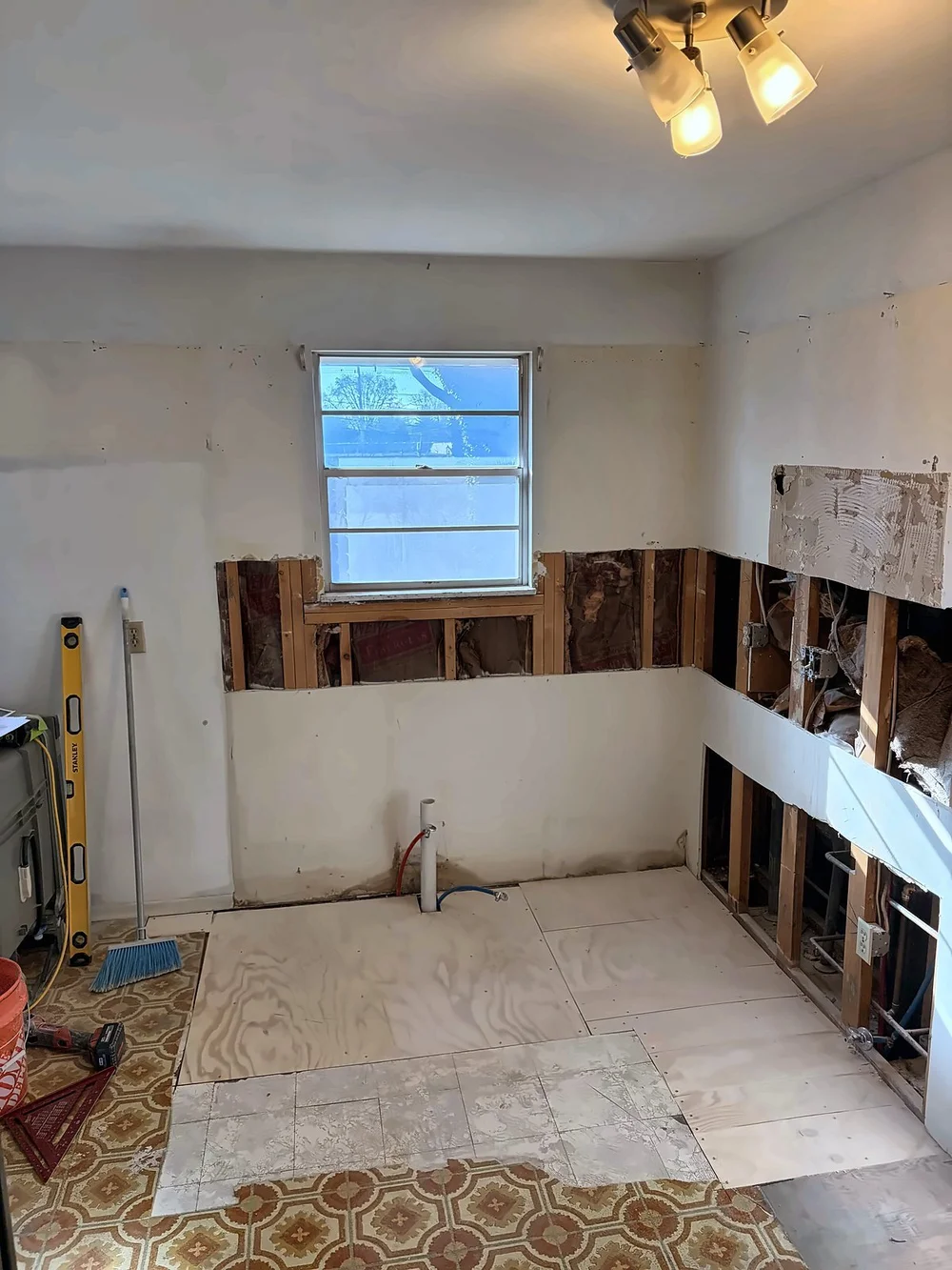 Kitchen during demolition with the drywall cut out below the window for cement board backsplash prep, bare plywood subfloor and old patterned vinyl at the edges